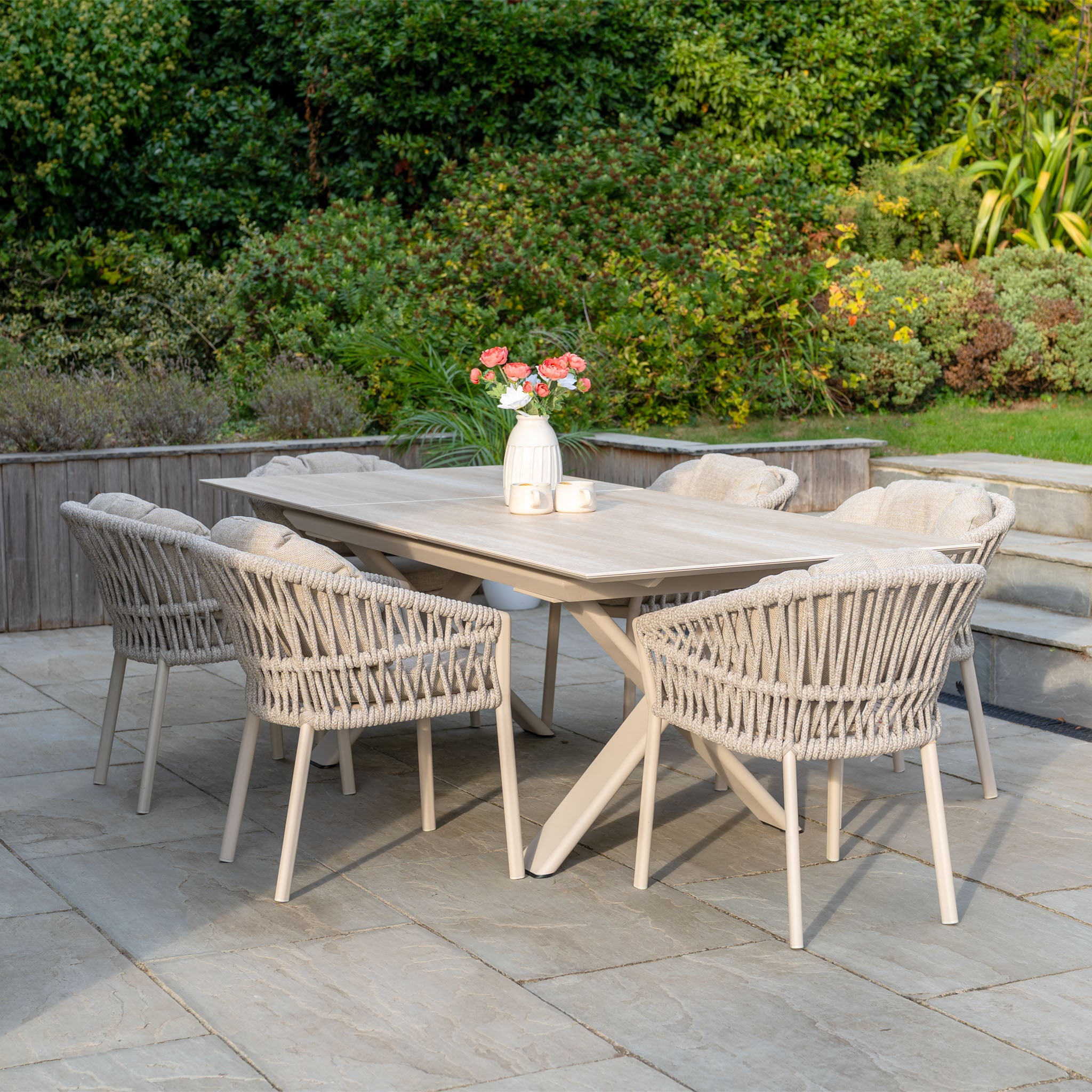 The Pello 6 Seat Dining Set with Albatross Extending Table in Fawn, shown with a vase of flowers and cups, is arranged on a stone patio surrounded by lush greenery.