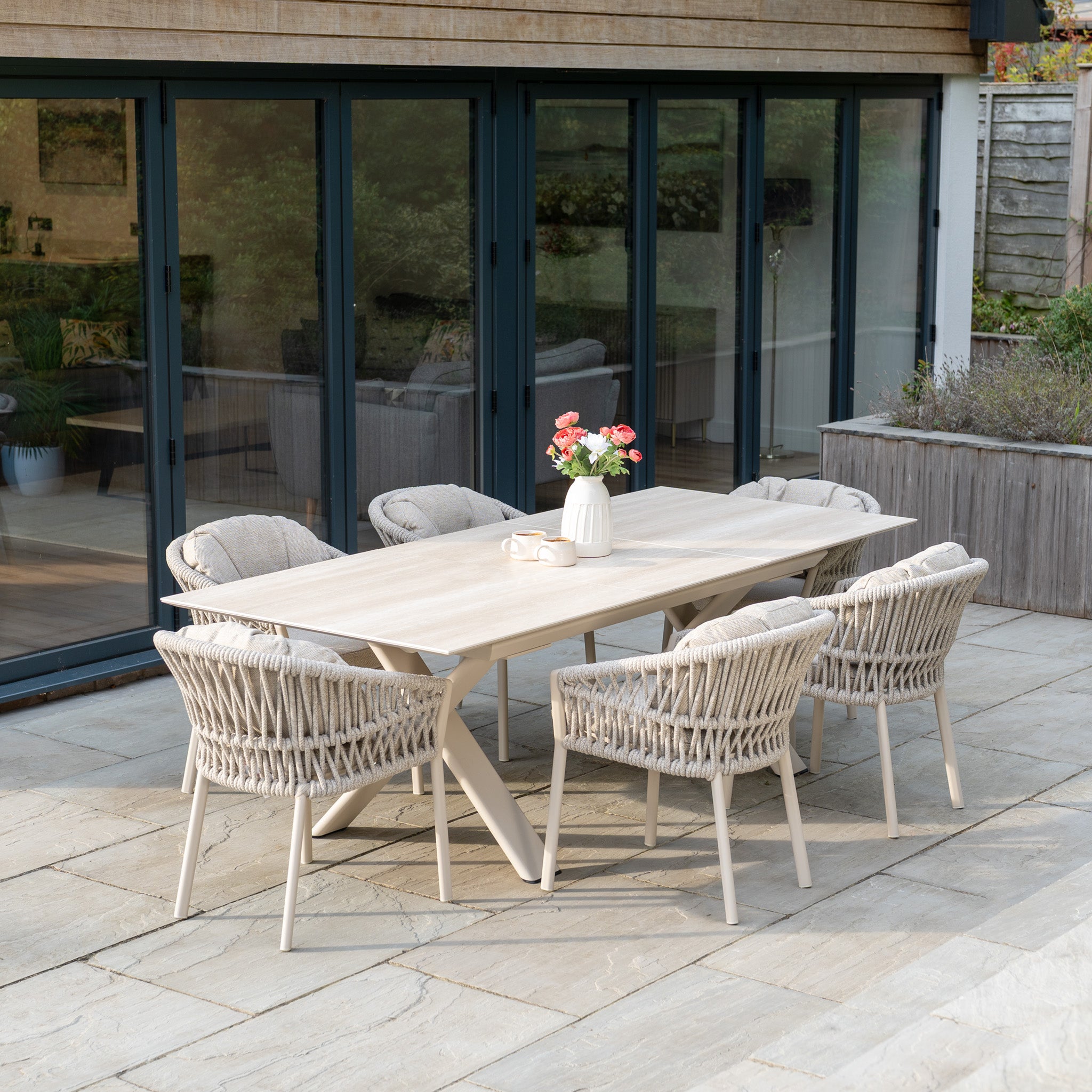 The Pello 6 Seat Dining Set with Albatross Extending Table in Fawn, featuring woven chairs, is arranged by glass doors. A vase of flowers and two mugs sit on the table, creating a perfect setting for outdoor gatherings.