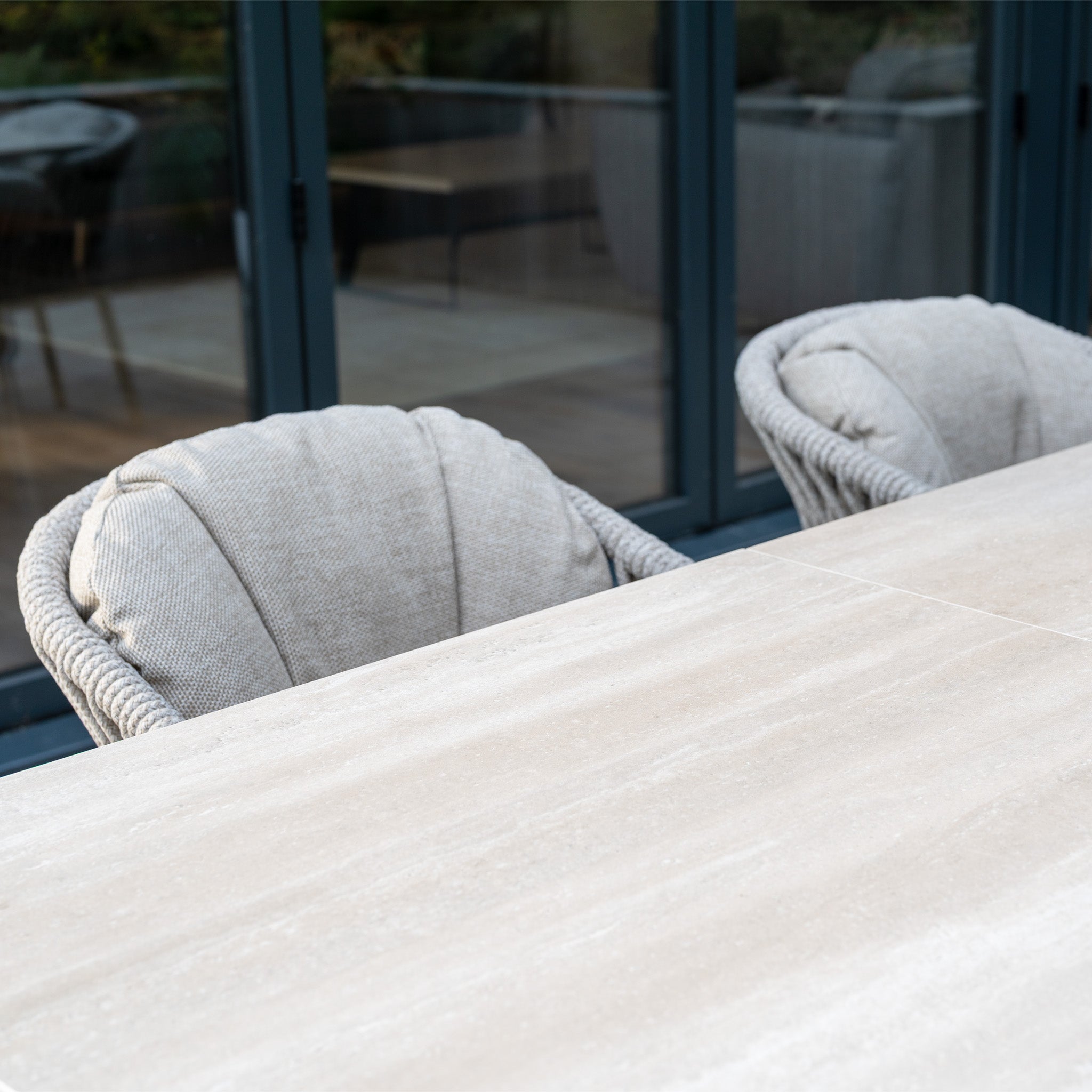 Two cushioned chairs are placed beside the Pello 6 Seat Dining Set with Albatross Extending Table in Fawn, positioned by glass doors that reflect the outdoor patio and chic dining furniture.