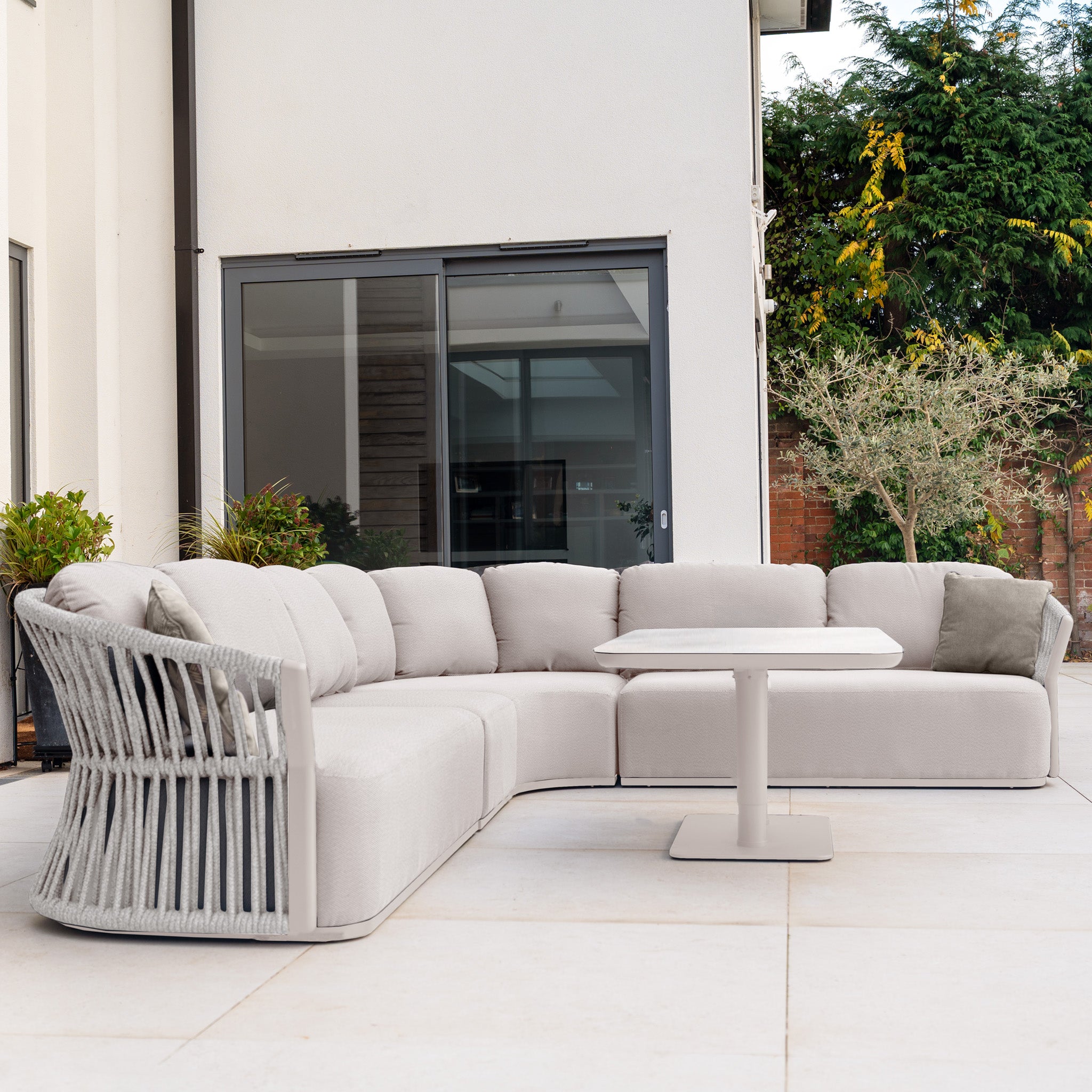 The Pello Deluxe Curved Sofa Set with Altura Rising Table in Fawn features quick dry garden cushions, set on a modern patio with plants and large glass doors in the background.