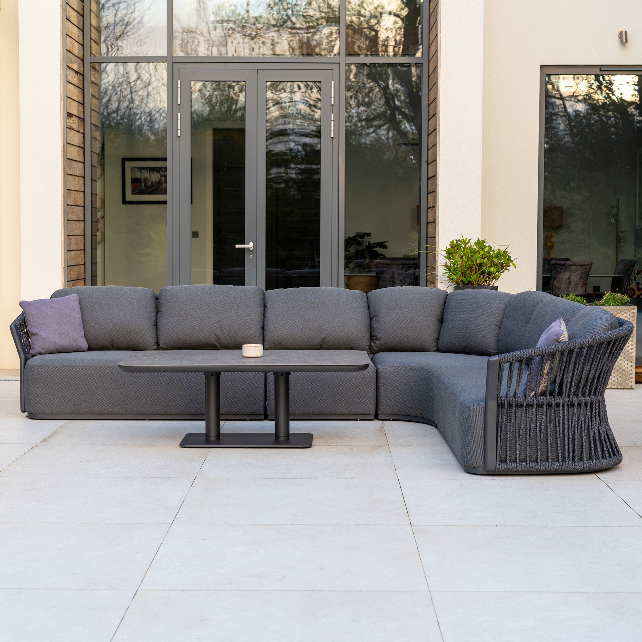 Modern outdoor patio with a Pello Deluxe Curved Sofa Set with Altura Rising Table in Grey and stylish cushions in front of large glass doors—an inviting space featuring contemporary garden furniture.