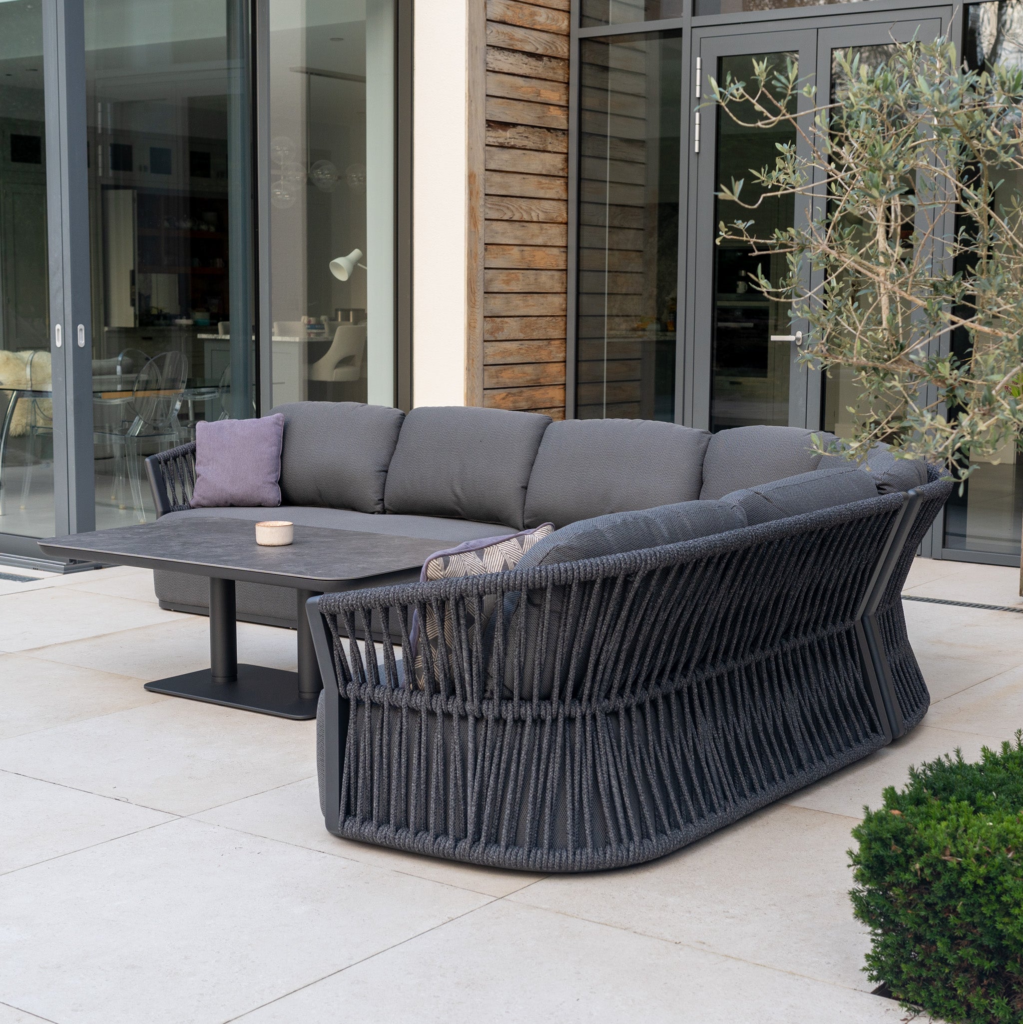 Modern patio with the Pello Deluxe Curved Sofa Set with Altura Rising Table in Grey—stylish garden furniture featuring gray cushions and a sleek table, set by large glass doors and lush plants.