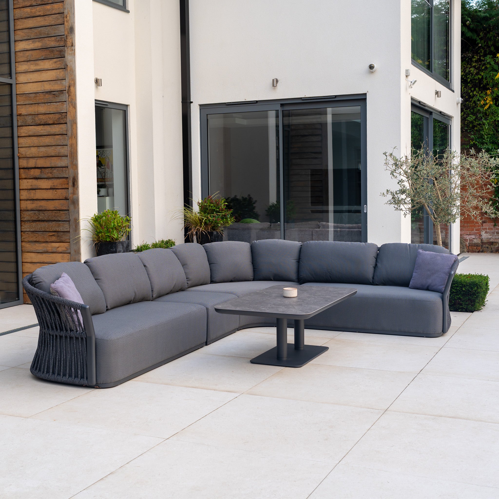 Modern outdoor patio featuring the Pello Deluxe Curved Sofa Set with Altura Rising Table in Grey and potted plants—perfect garden furniture for relaxing outdoors.