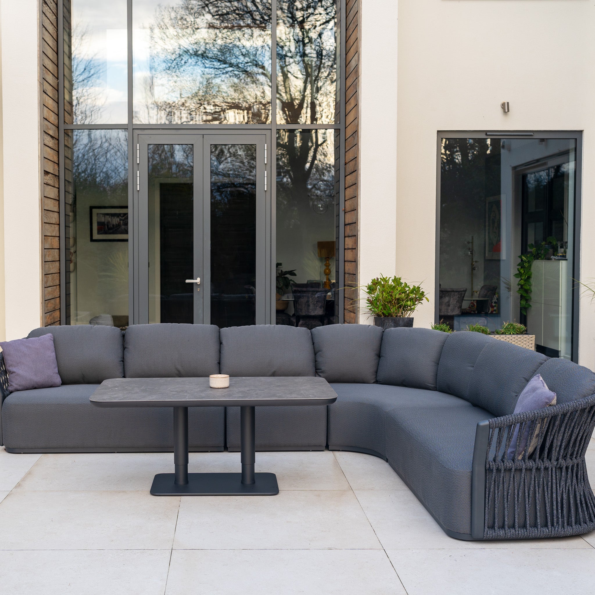 The Pello Deluxe Curved Sofa Set with Altura Rising Table in Grey is a stylish outdoor furniture set with cushions and a versatile table, perfect for your patio or garden.