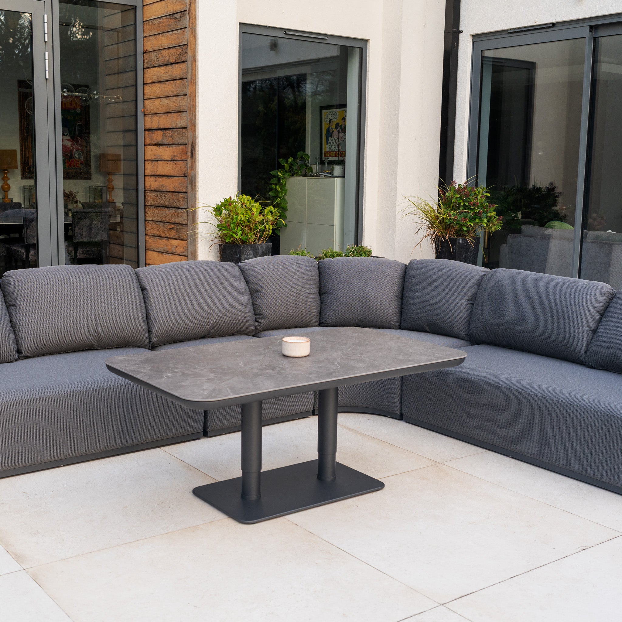 The Pello Deluxe Curved Sofa Set with Altura Rising Table in Grey creates a stylish, inviting outdoor space—perfect for relaxing on the patio near large windows and potted plants.