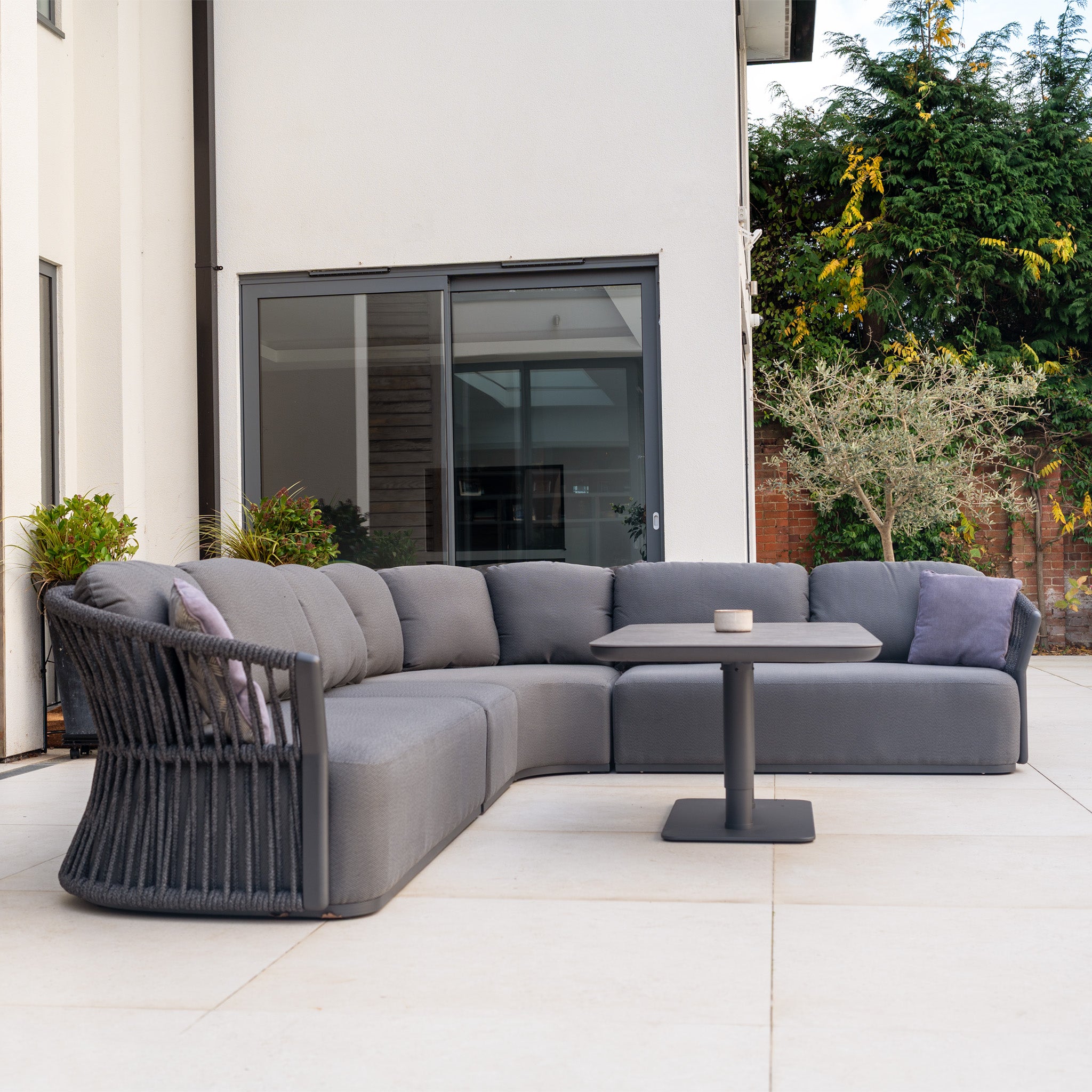 Modern outdoor patio featuring the Pello Deluxe Curved Sofa Set with Altura Rising Table in Grey, plush cushions, and lush greenery by a white house—ideal garden furniture for relaxing or entertaining.