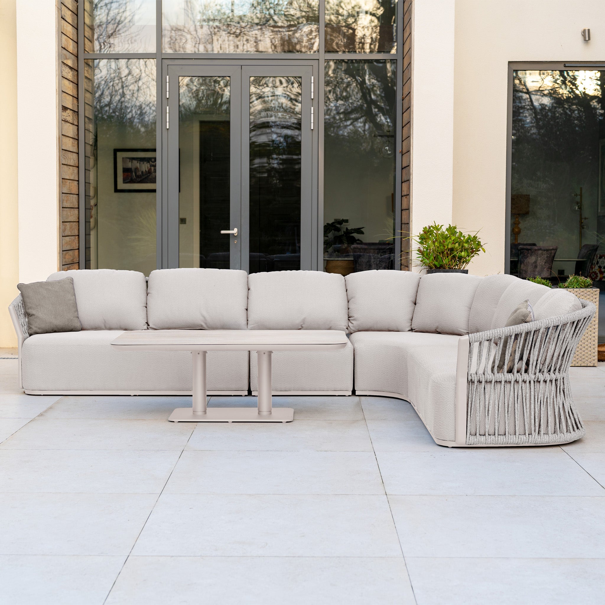 The Pello Deluxe Curved Sofa Set with Altura Rising Table in Fawn features quick-dry garden cushions, shown on a patio before glass doors and windows.
