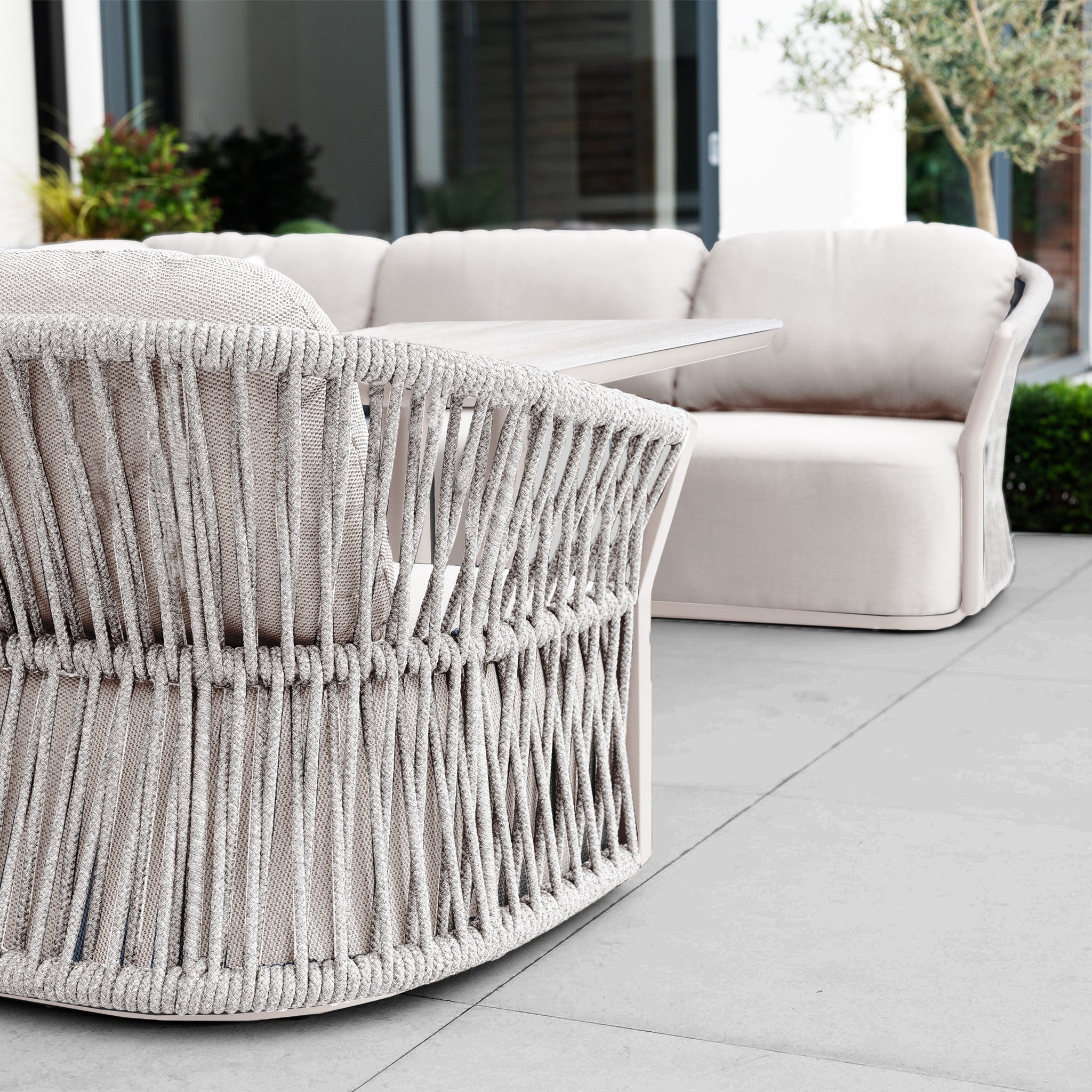 Outdoor patio with the Pello U-Shape Corner Sofa Set and Altura Rising Table in Fawn, plus light beige woven chairs arranged on tiled flooring.