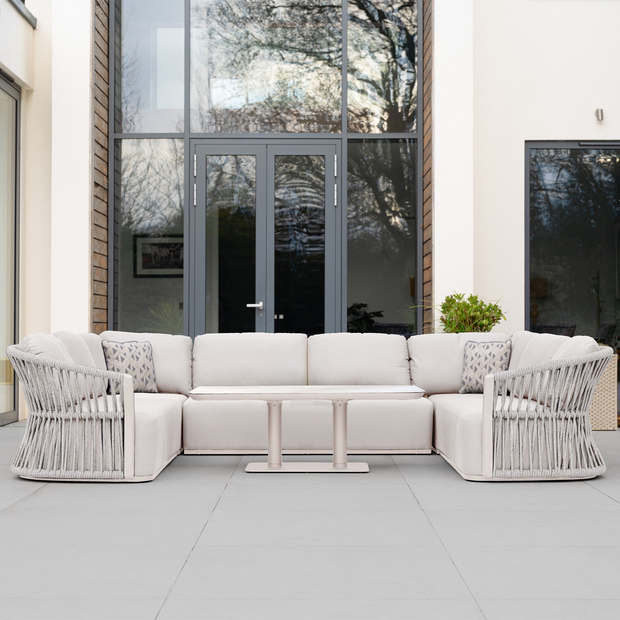 A modern patio with the Pello U-Shape Corner Sofa Set in Fawn and Altura Rising Table, styled with decorative pillows, elegantly arranged before glass doors.