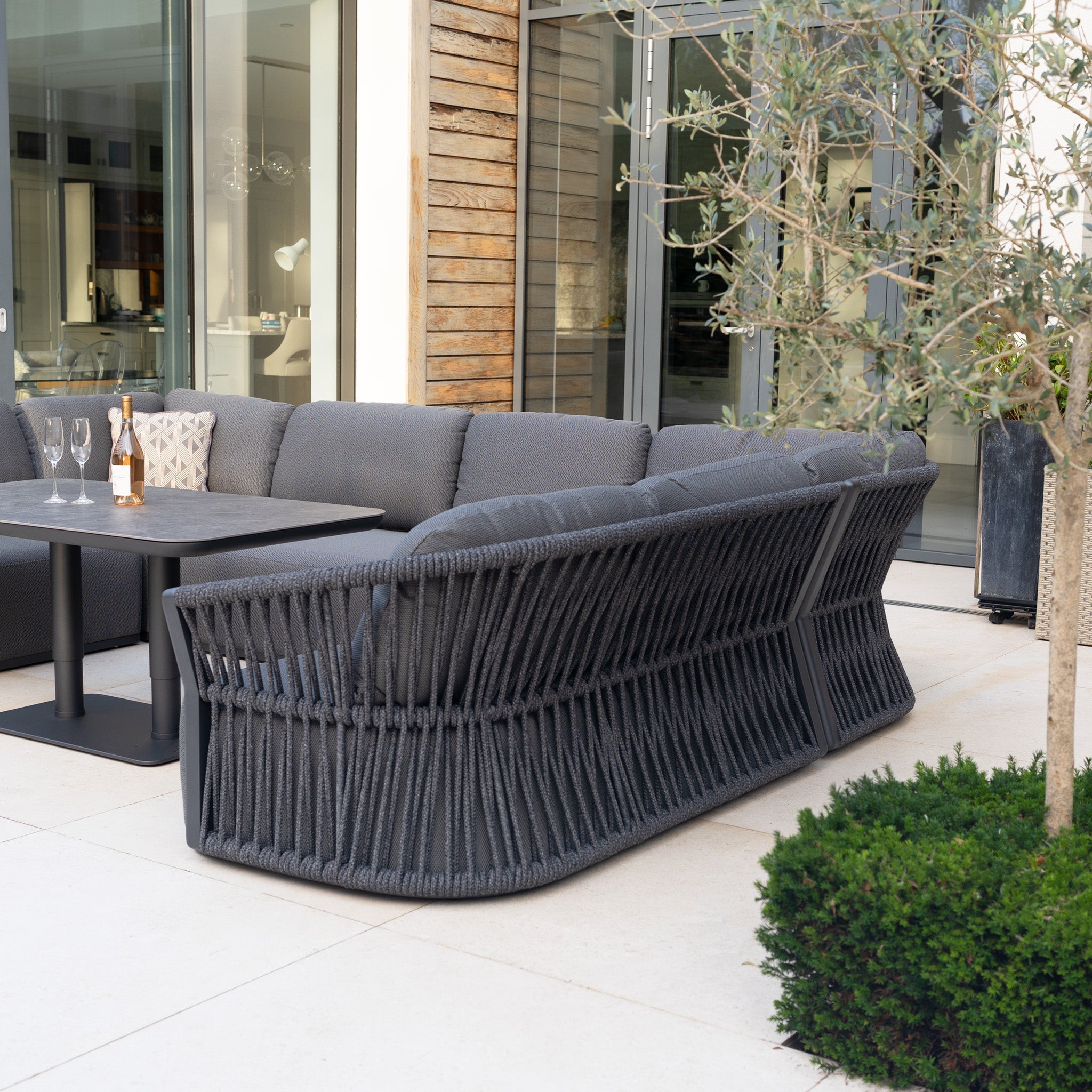 Outdoor patio featuring the Pello U-Shape Corner Sofa Set with Altura Rising Table in Grey, complemented by wine glasses on the table and a nearby potted plant.