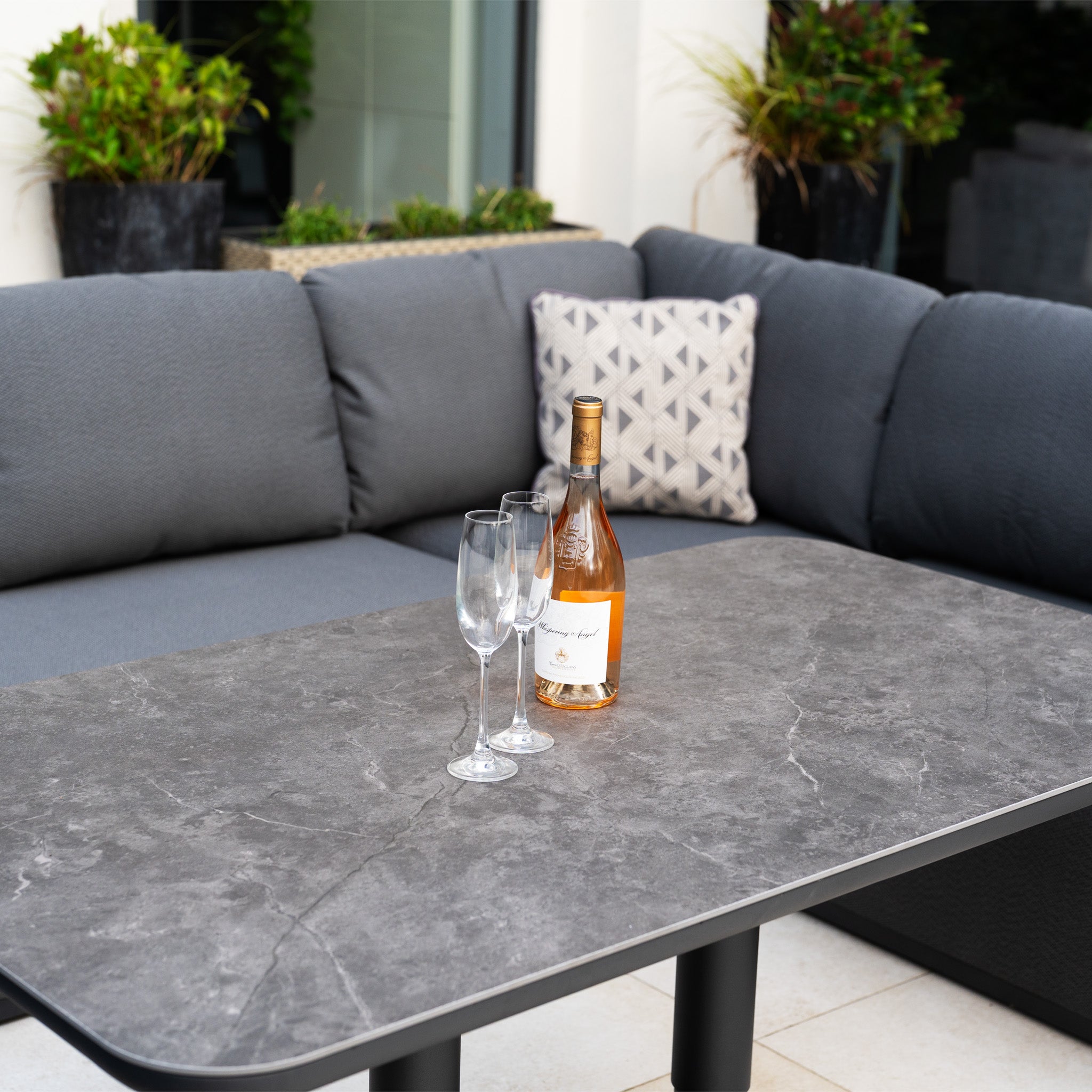 A bottle of rosé wine and three empty glasses rest on a gray stone table, with the Pello U-Shape Corner Sofa Set with Altura Rising Table in Grey featured stylishly in the background.