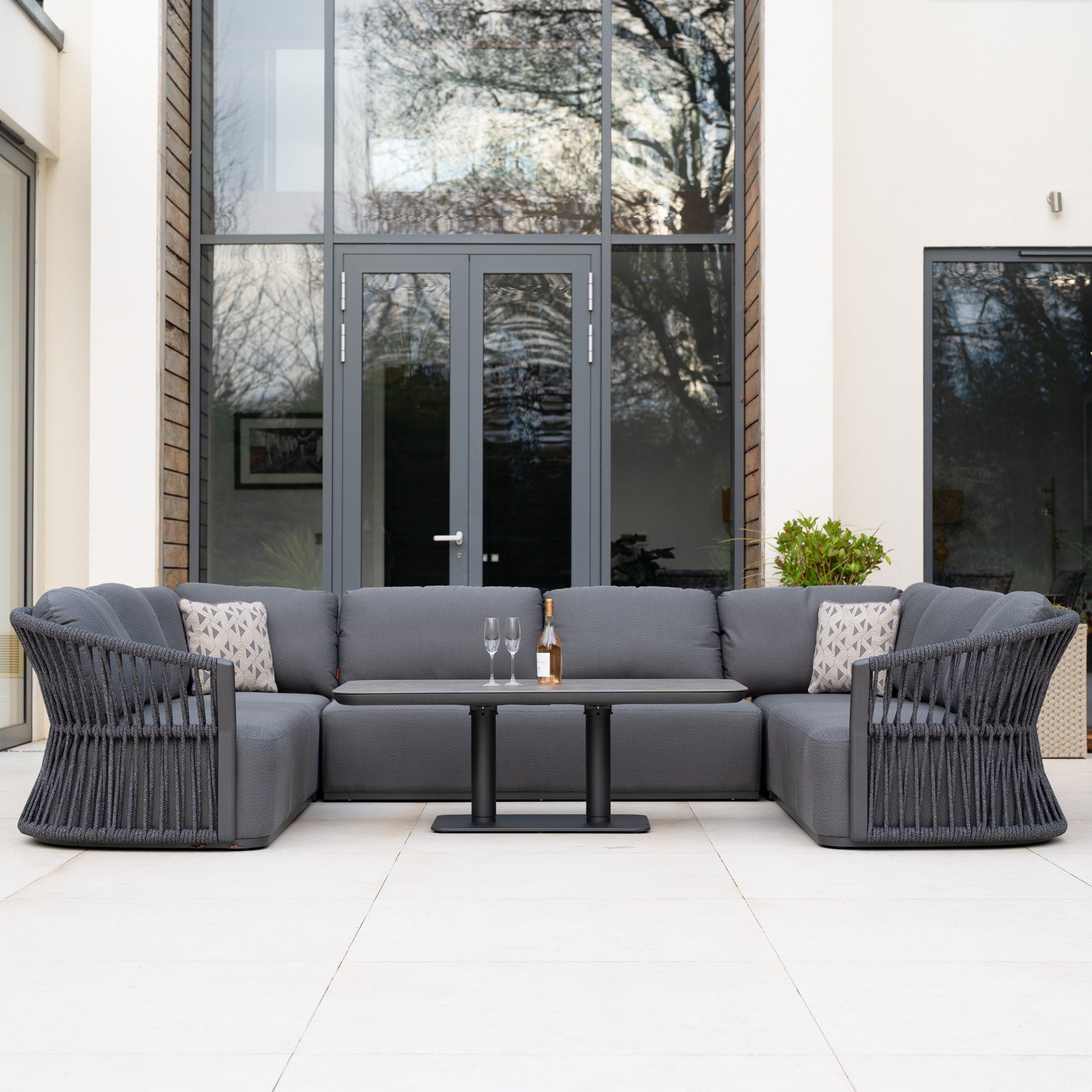 The Pello U-Shape Corner Sofa Set with Altura Rising Table in Grey is arranged outdoors, featuring a modern curved sofa and table placed before expansive glass doors.