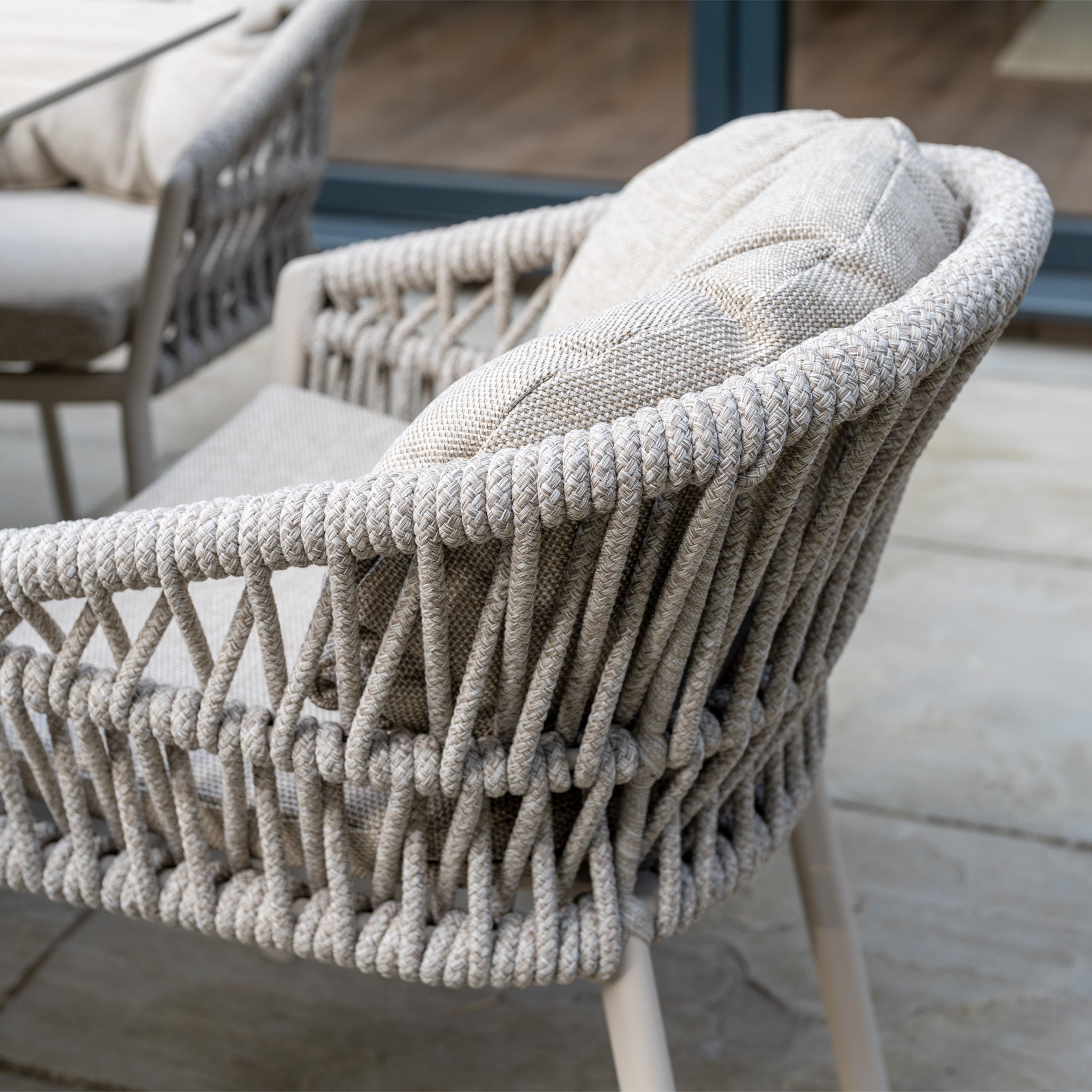 Close-up of the Pello 8 Seat Oval Dining Set with Ceramic Table in Fawn, featuring a woven outdoor chair with beige cushions, all designed for weather resistance and displayed on a stone patio.