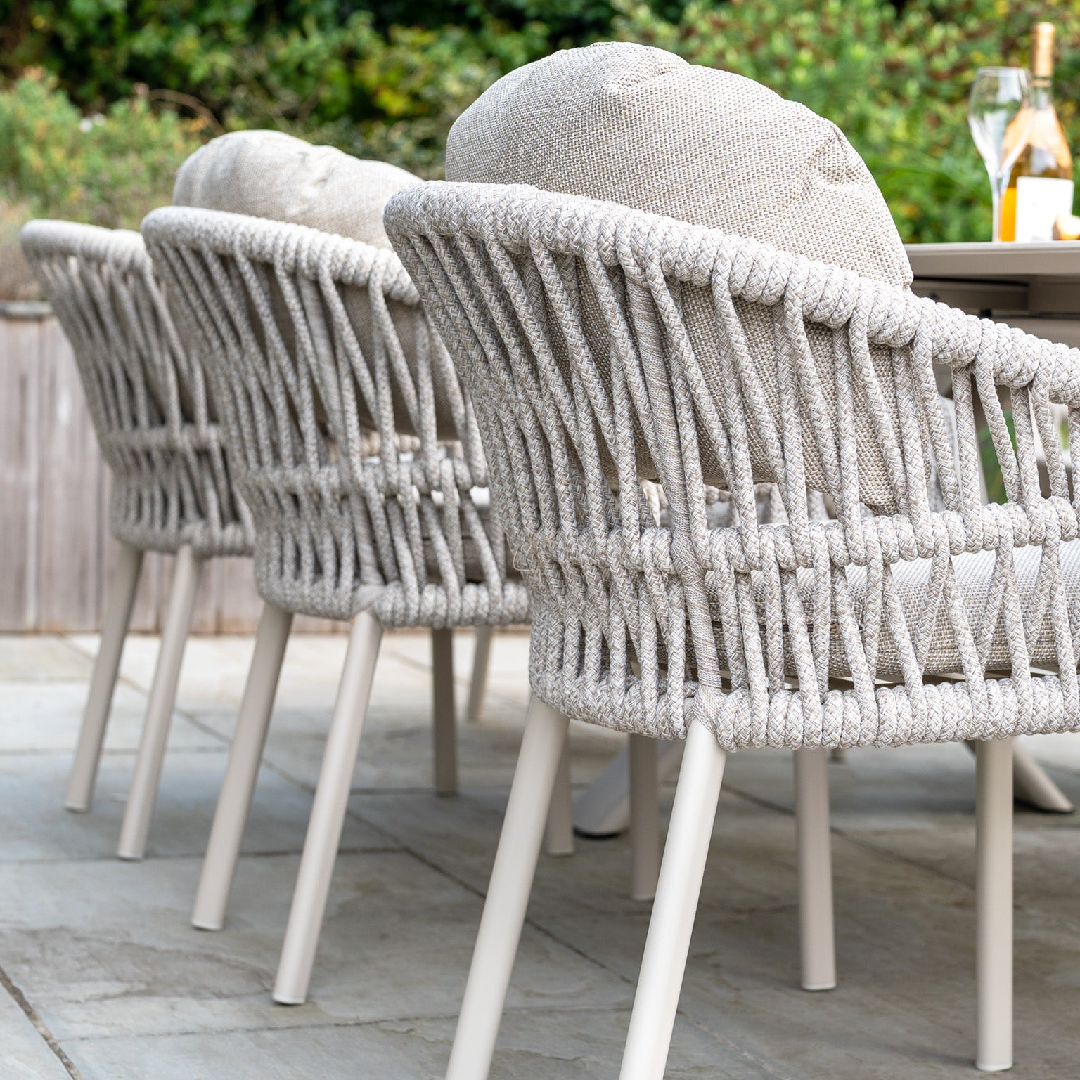 The Pello 6 Seat Oval Dining Set with Ceramic Table in Fawn features stylish, weather-resistant chairs and a sleek ceramic table on a stone patio, providing both durability and elegance amid lush greenery.