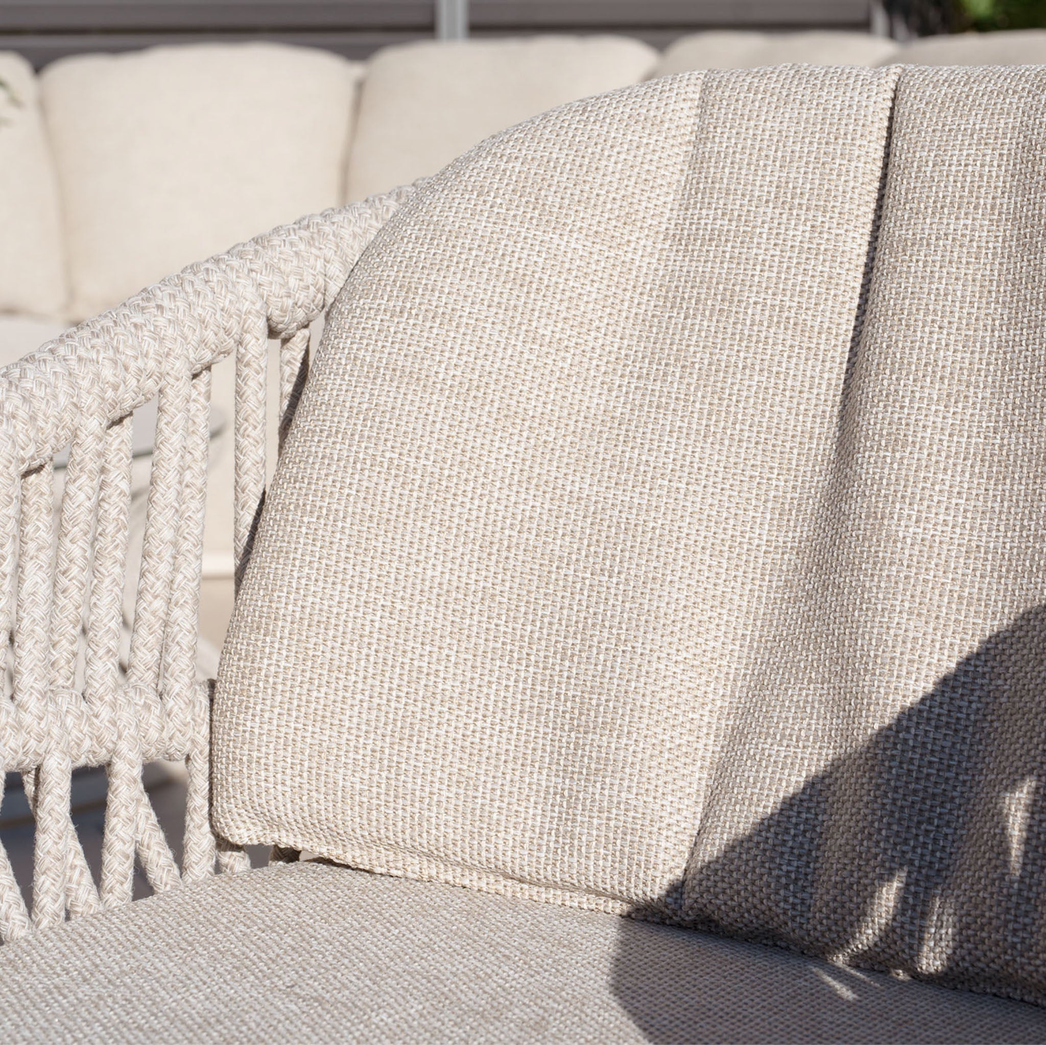Close-up of the Pello 8 Seat Extending Dining Set's beige woven outdoor chair in Fawn, featuring a solution-dyed olefin cushion and sunlight casting shadows across the ceramic table and seat.