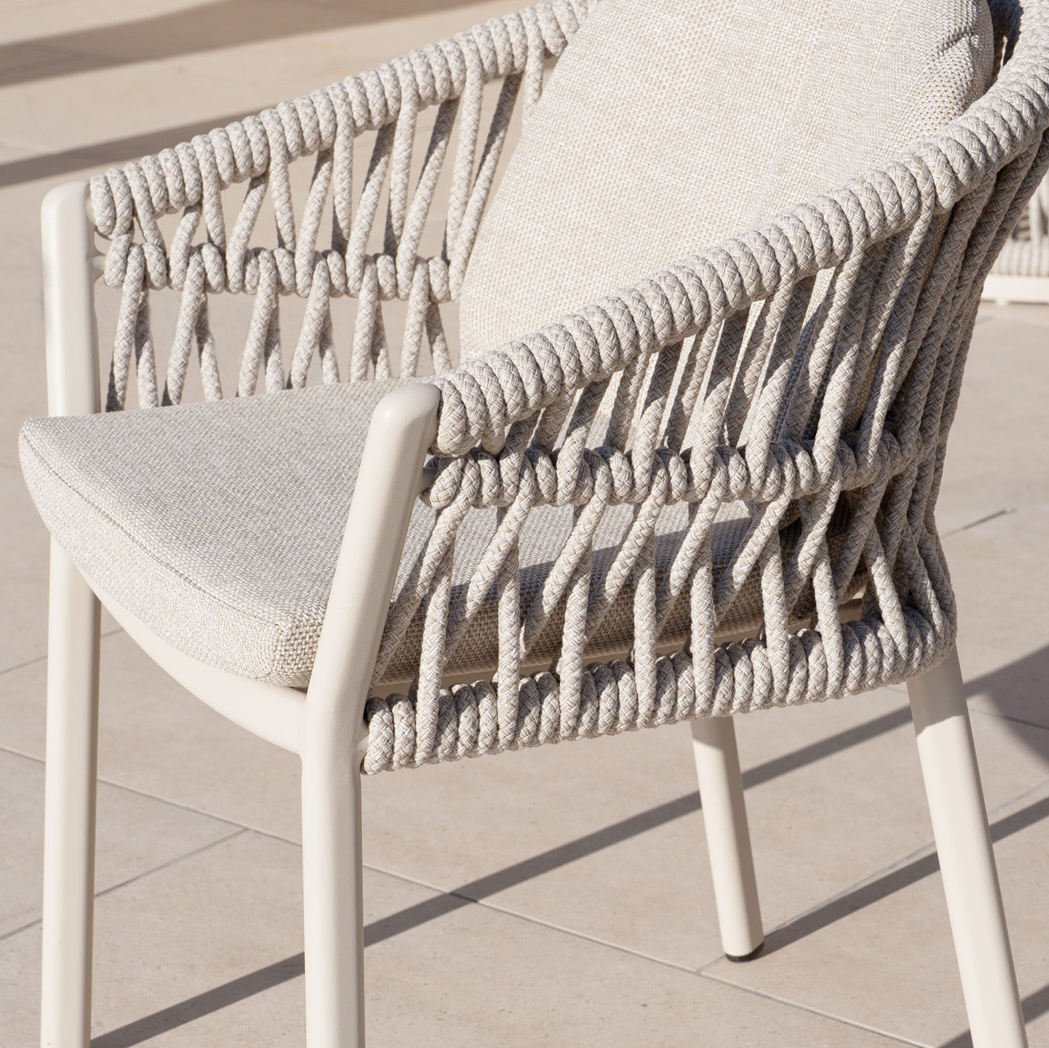 A beige chair with modern woven detailing and a cushion sits on a sunny tiled patio—ideal for outdoor gatherings with your Pello 10 Seat Extending Dining Set with Ceramic Table in Fawn.