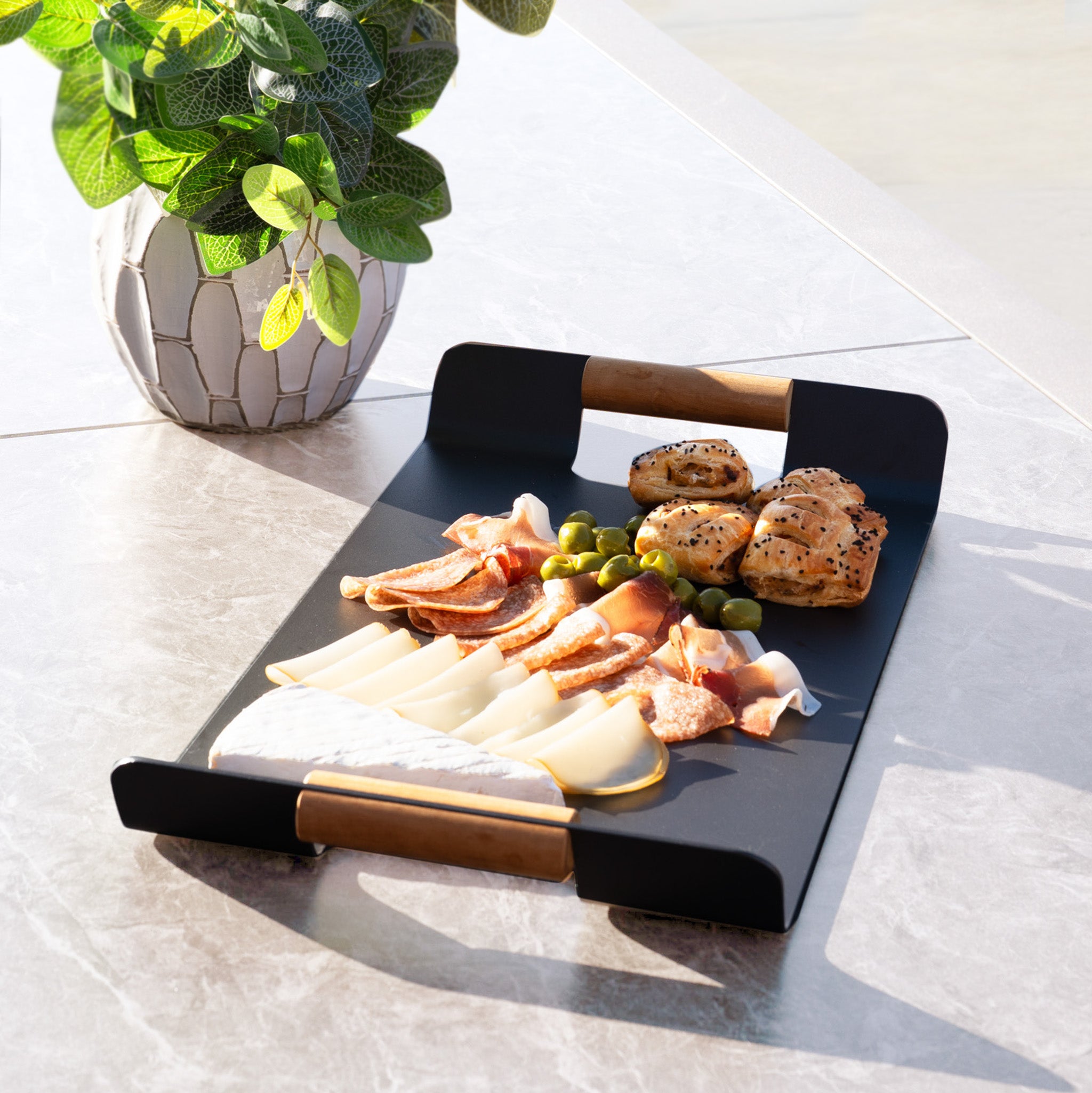 A charcuterie tray with meats, cheeses, olives, and bread sits on a marble table beside a potted plant, perfect for outdoor dining with the Pello 8 Seat Extending Dining Set with Ceramic Table in Fawn.