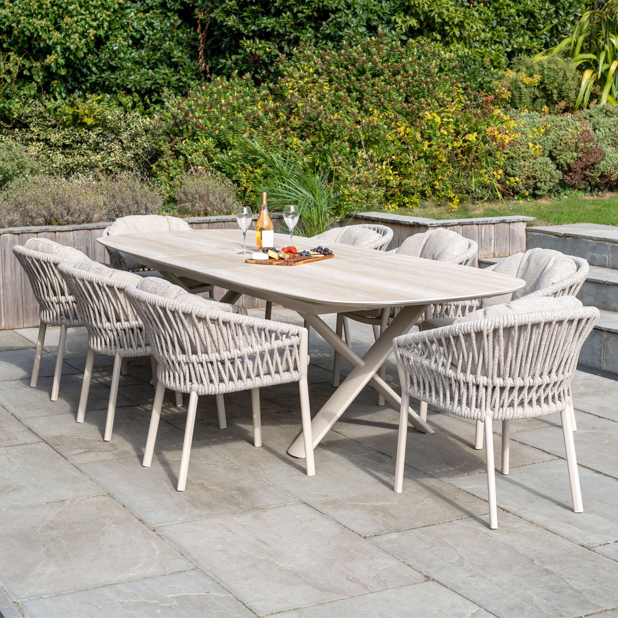 The Pello 8 Seat Oval Dining Set with Albatross Extending Table in Fawn features eight woven chairs and a sturdy extending table, stylishly set with drinks and snacks on a stone terrace.