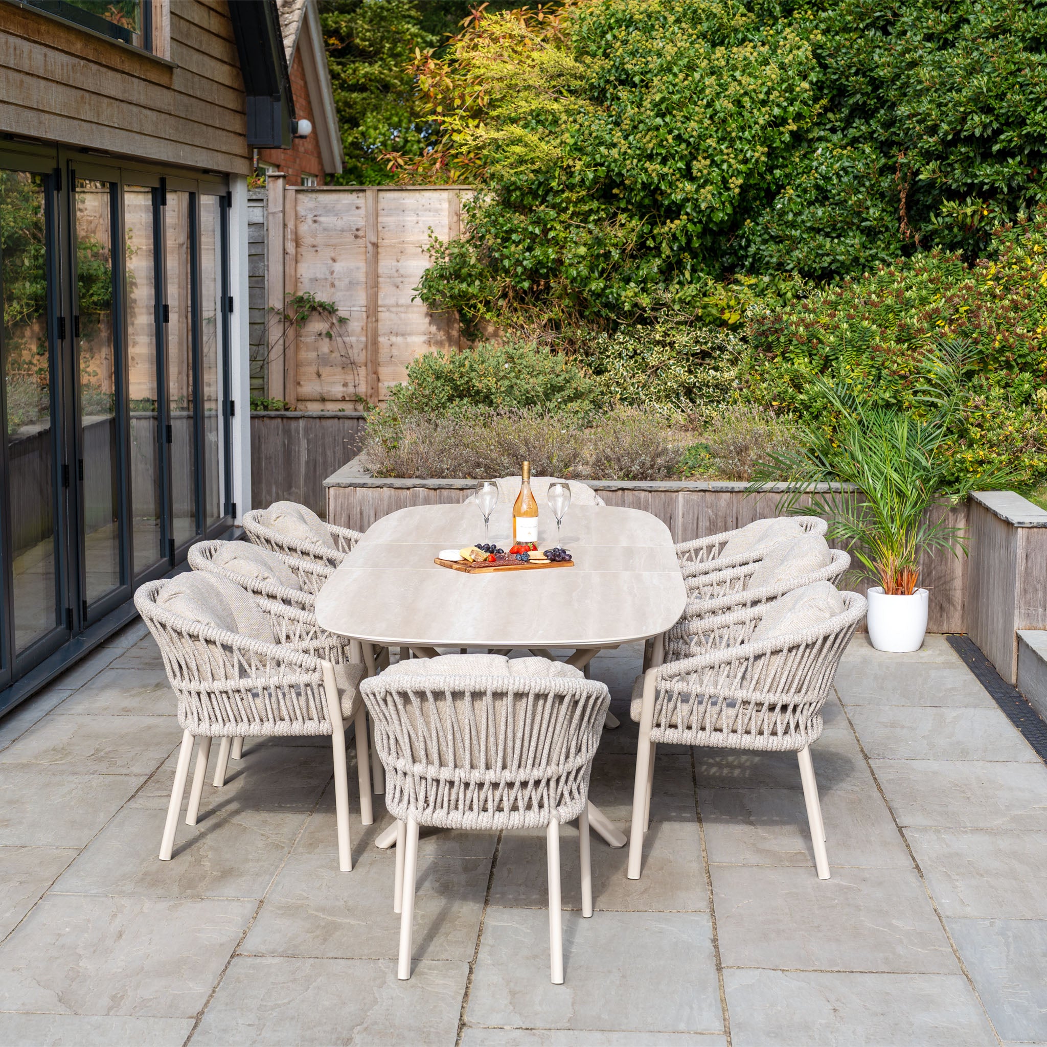 The Pello 8 Seat Oval Dining Set with Albatross Extending Table in Fawn features eight woven chairs and a spacious, durable table—ideal garden furniture for outdoor dining on a patio surrounded by greenery.