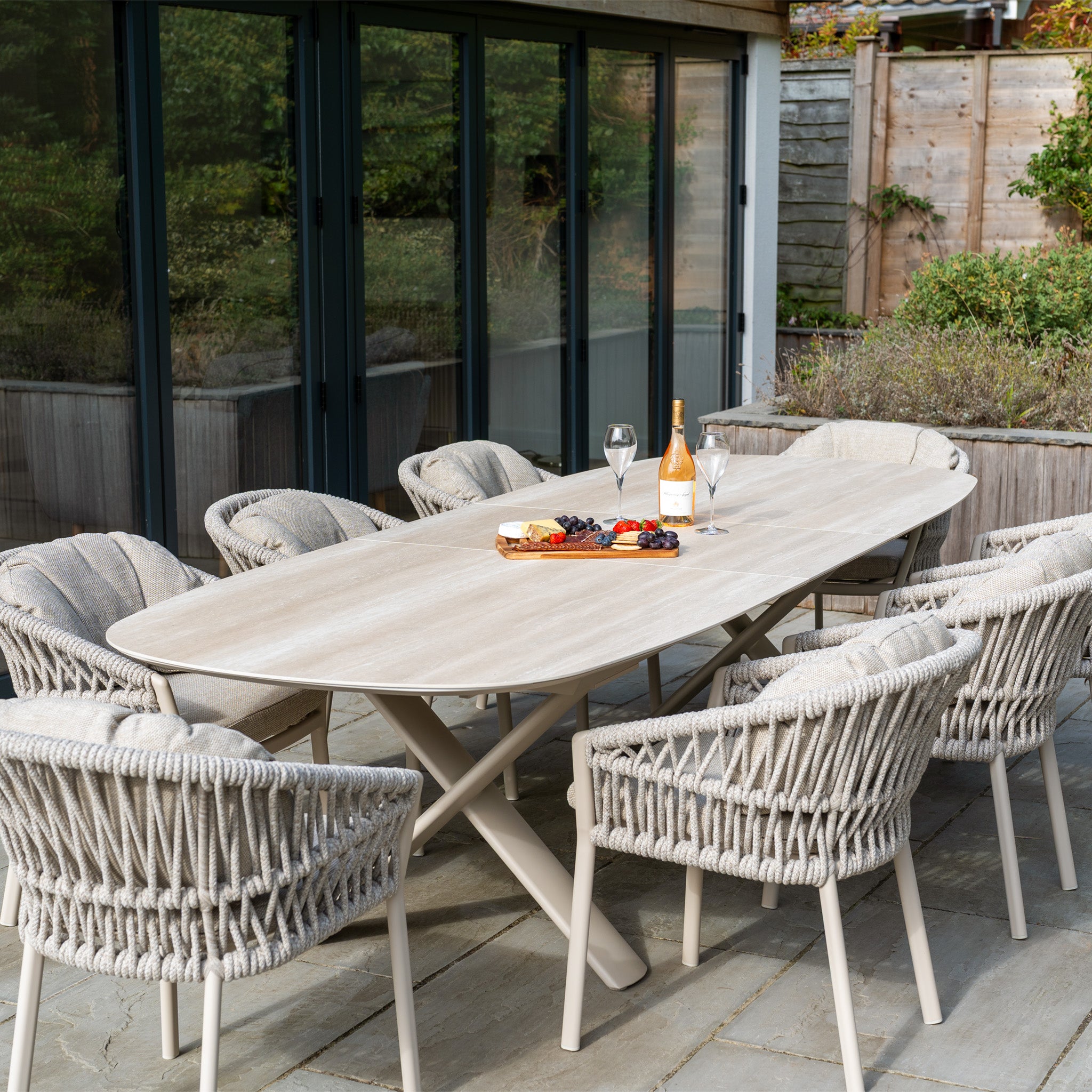 The Pello 8 Seat Oval Dining Set with Albatross Extending Table in Fawn seats eight and is ideal for al fresco gatherings, shown here with wine, glasses, cheese, and fruit on a stone patio by glass doors. Durable garden furniture for outdoor dining.