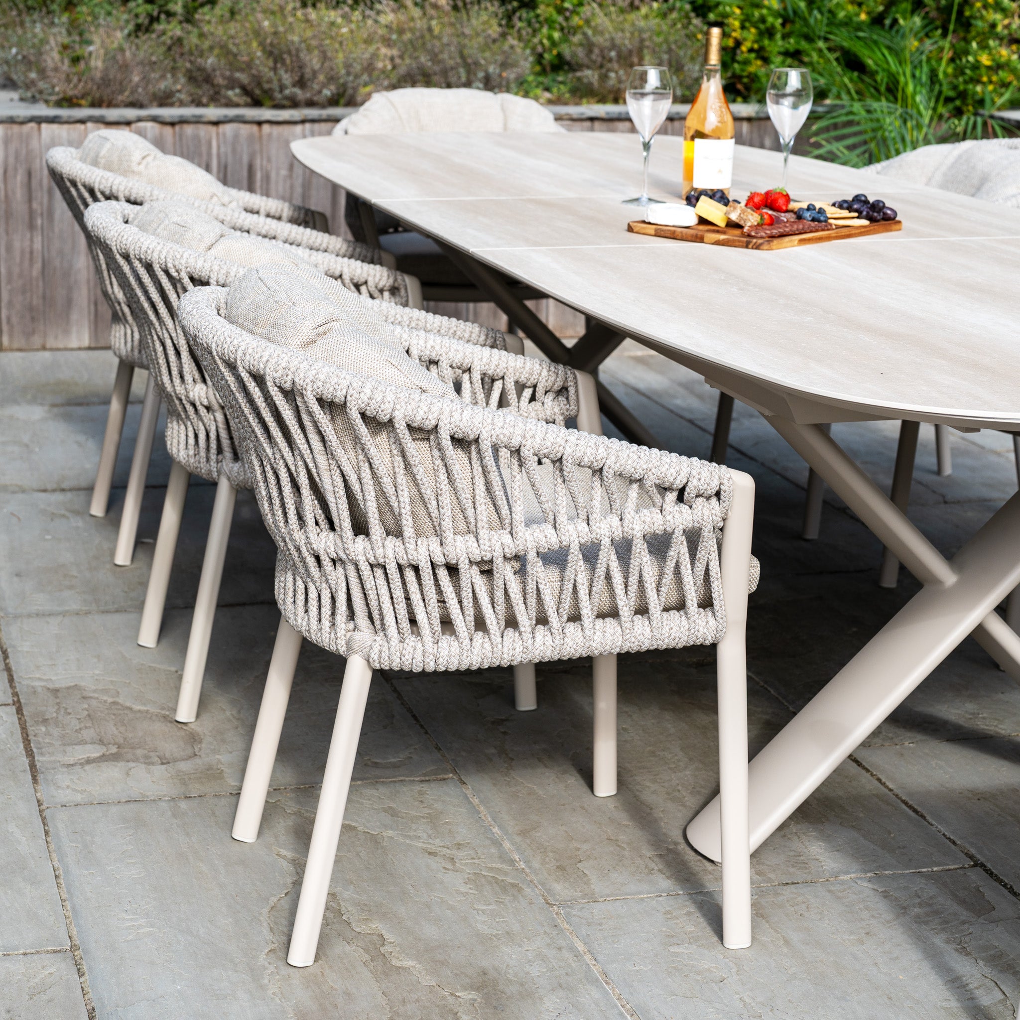 The Pello 8 Seat Oval Dining Set with Albatross Extending Table in Fawn includes a sturdy garden table, woven chairs, wine glasses, a bottle, and a cheese board with fruit for stylish outdoor dining.