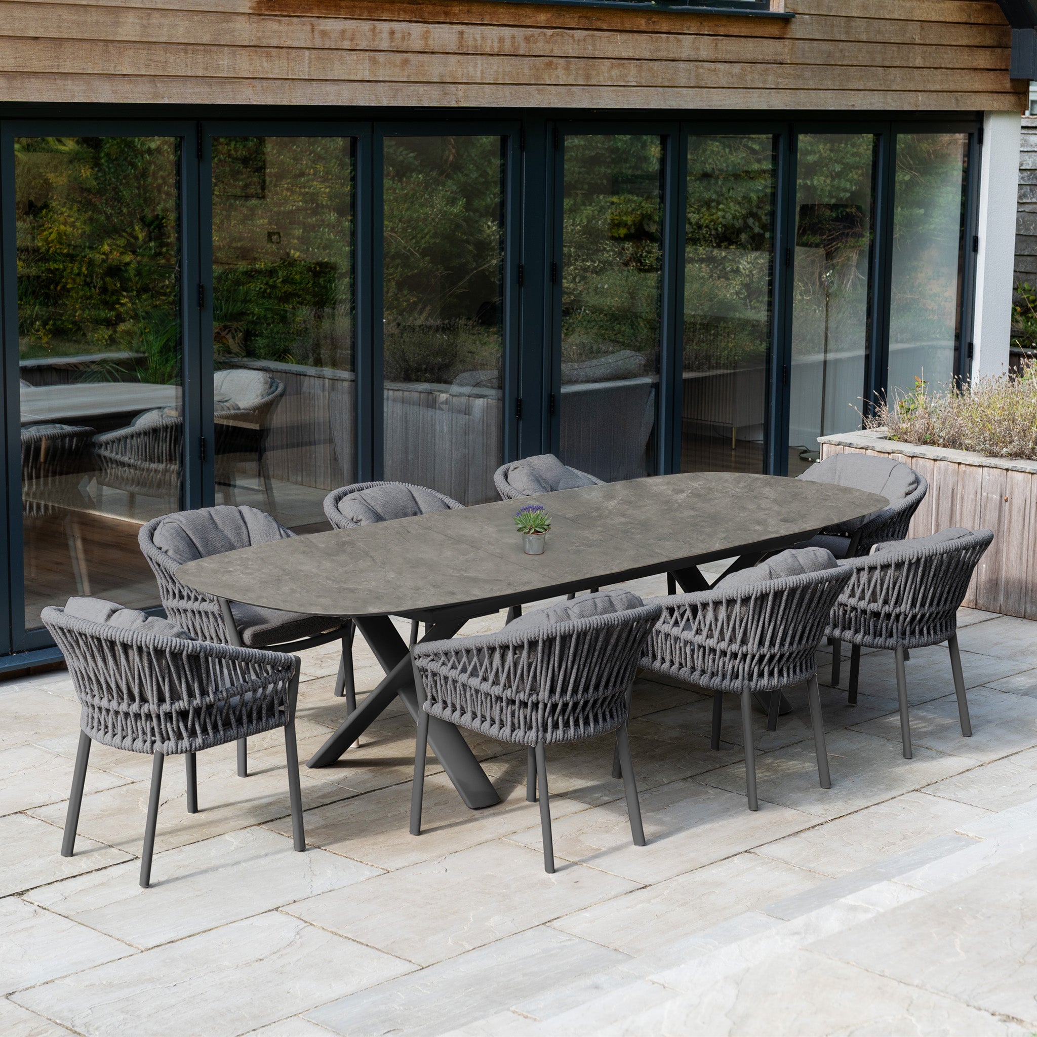 The Pello 8 Seat Oval Dining Set with Albatross Extending Table in Grey features eight woven chairs, perfect for outdoor dining on a stone patio in front of spacious glass doors.