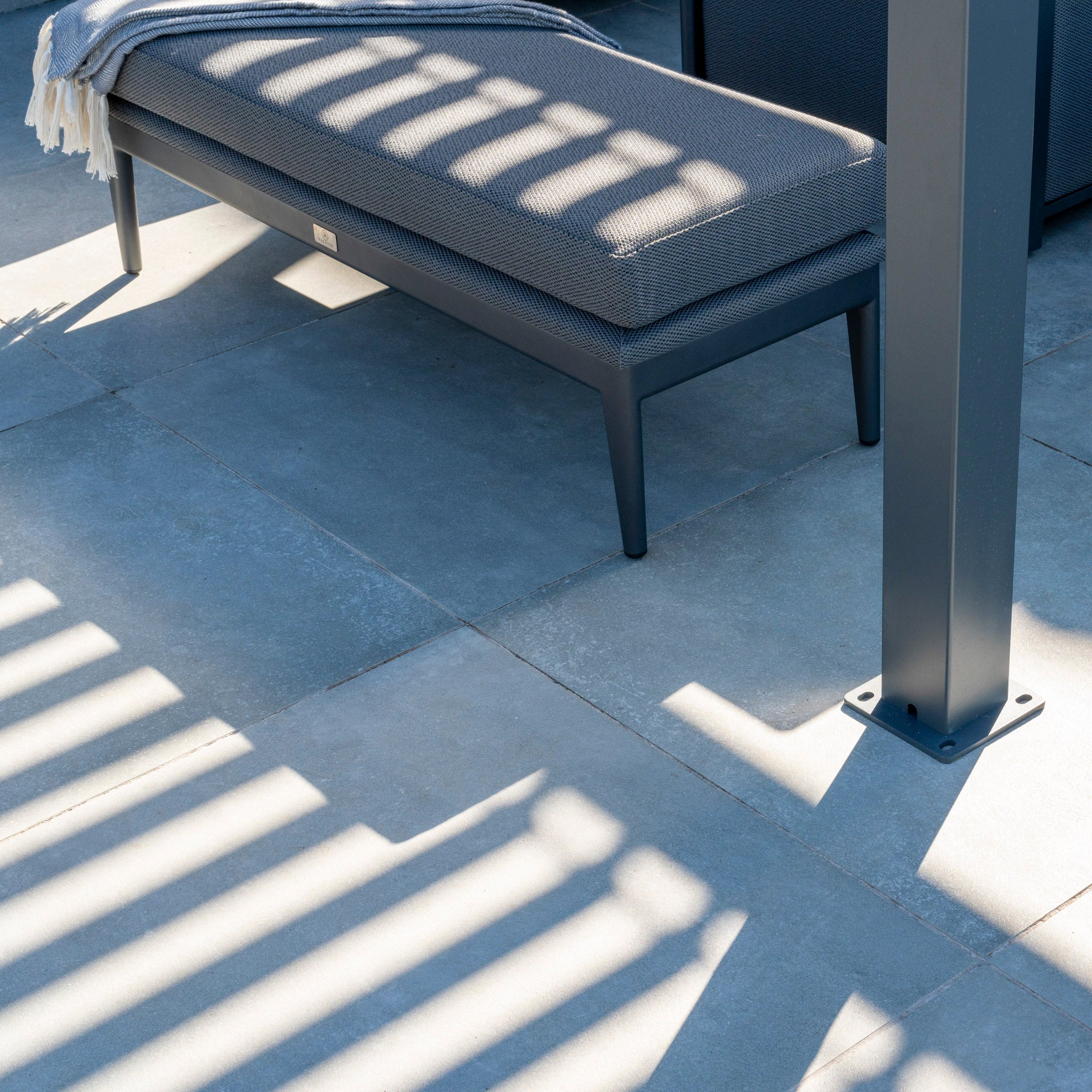 On a stone patio, an outdoor bench catches striped shadows from the PergoSTET Lite 3m x 3m Metal Square Pergola in Grey overhead, boasting a sleek waterproof roof and built-in drainage. A draped blanket adds warmth and comfort to this sophisticated setting.