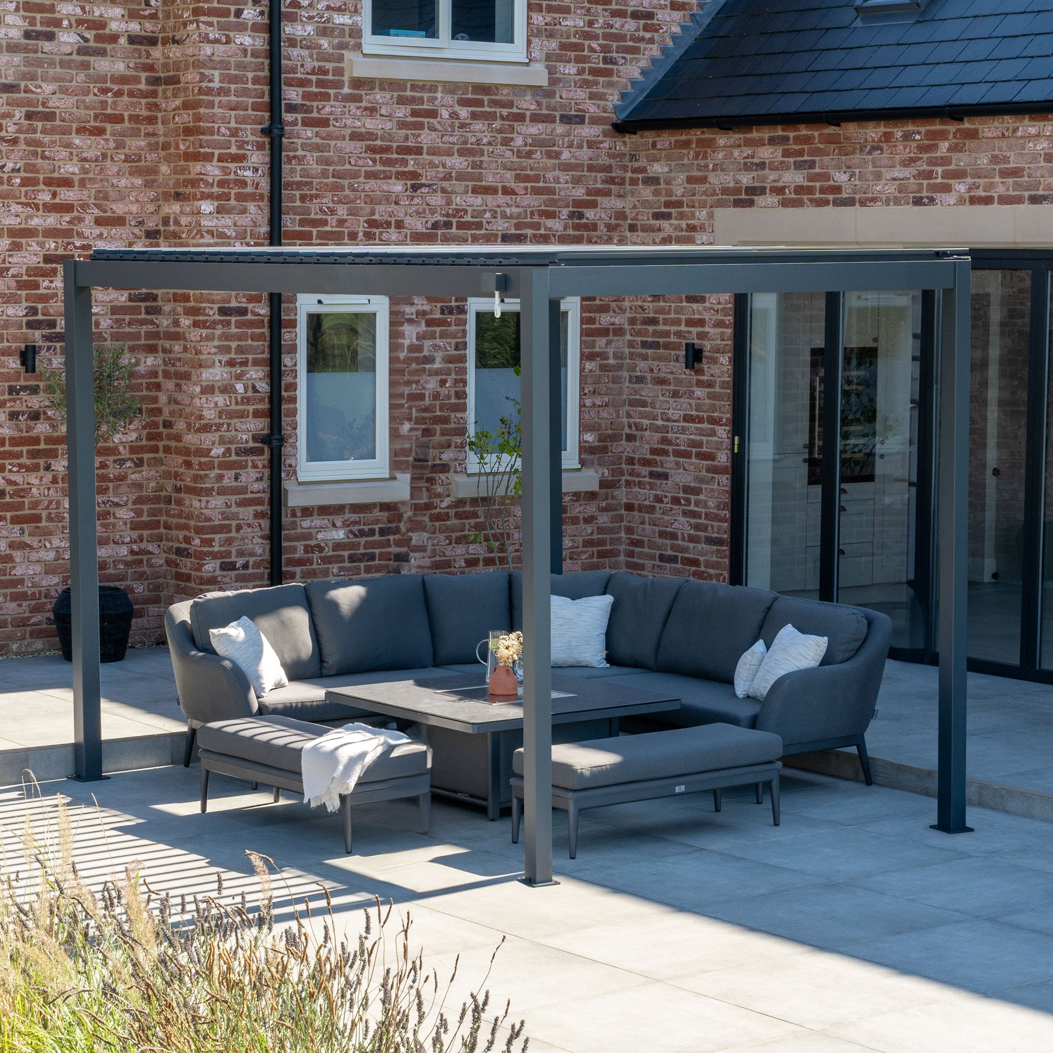 A sleek outdoor patio features a gray sectional sofa under the PergoSTET Lite 3m x 4m Metal Rectangular Pergola in Grey, complete with a built-in drainage system. The elegant design harmonizes beautifully with the brick house backdrop.