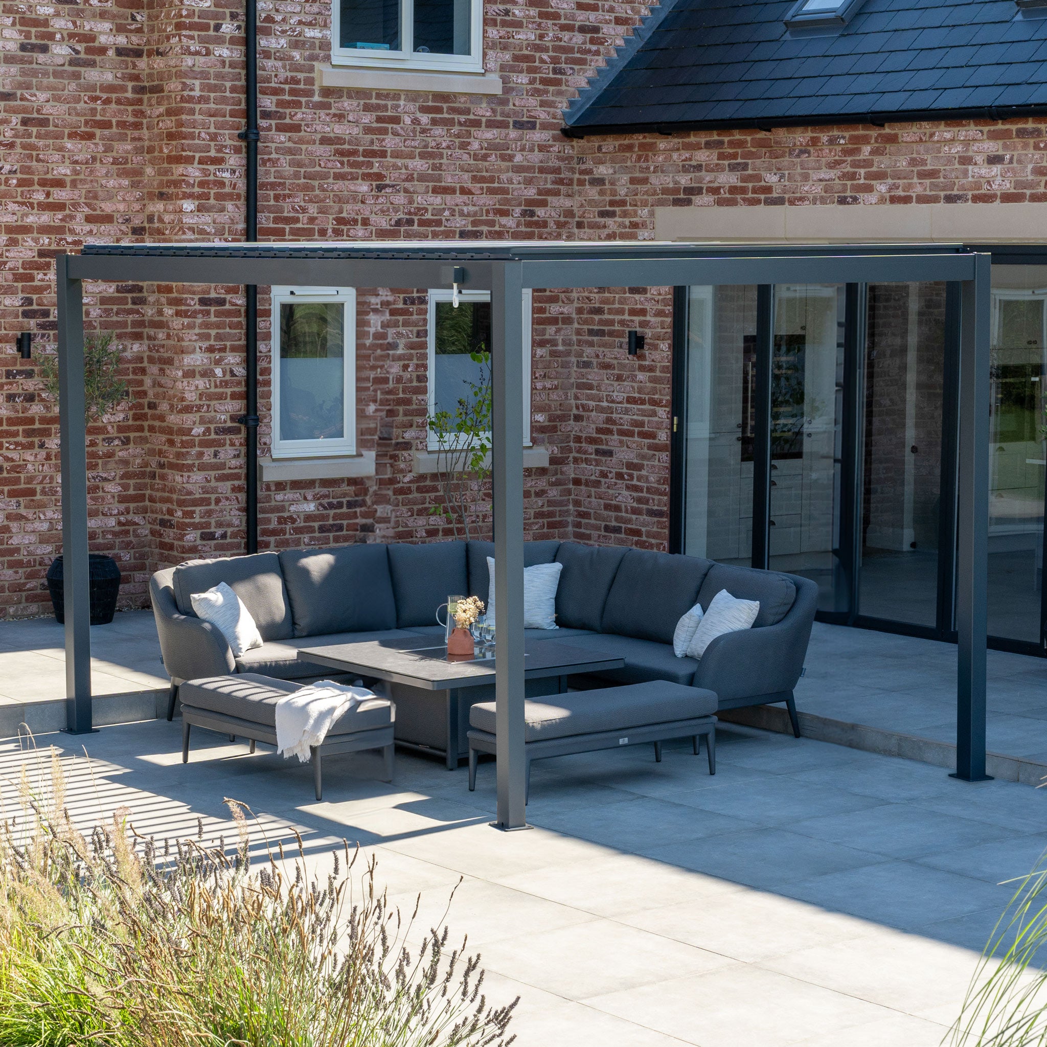 A modern patio features a gray sectional sofa under the sleek PergoSTET Lite 3m x 4m Metal Rectangular Pergola in Grey, adjacent to a brick house with large glass doors. The pergola's built-in drainage system provides comfort and style in any weather.