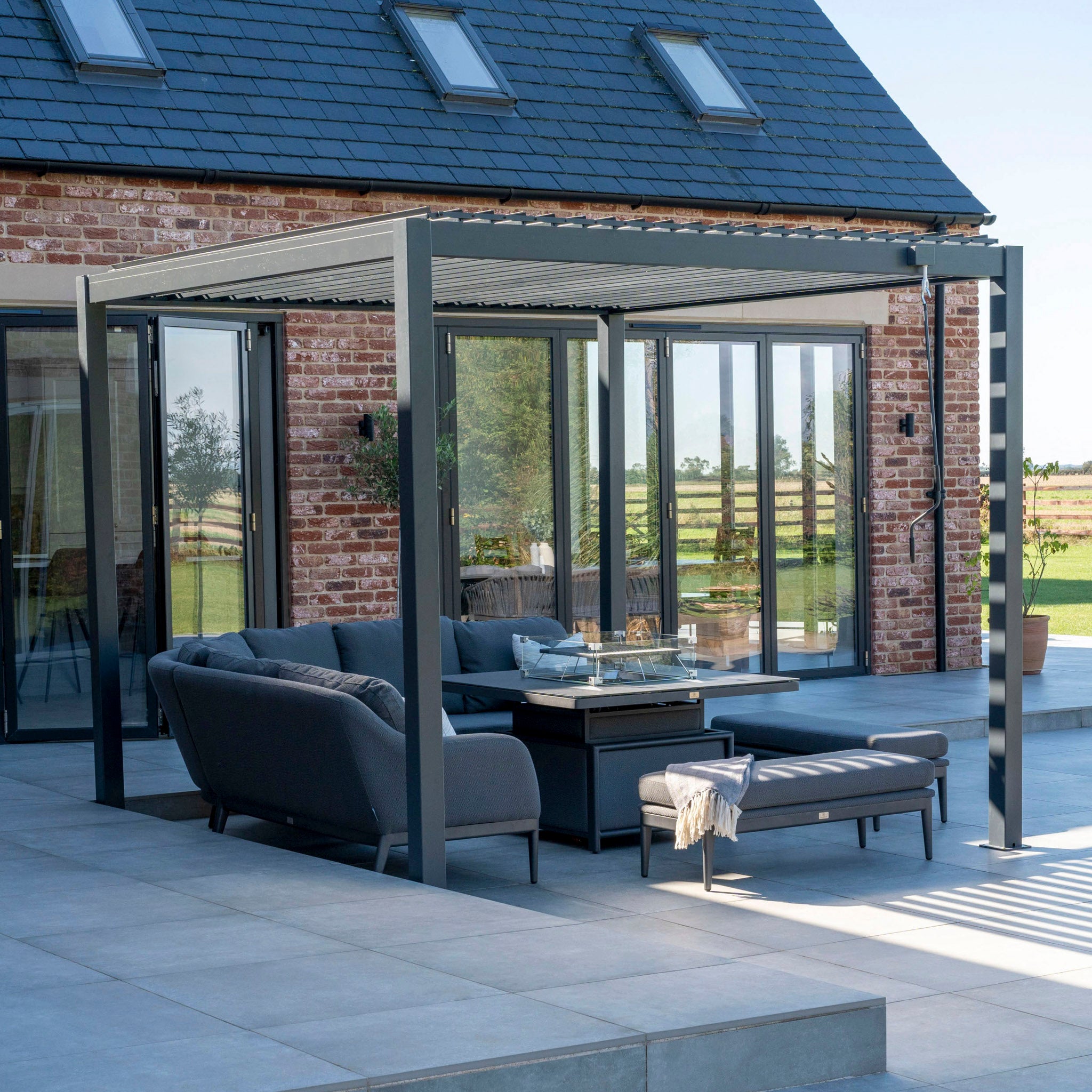 A modern patio showcases gray sofas and a coffee table under the PergoSTET Lite 3m x 3m Metal Square Pergola in Grey, complete with a built-in drainage system, all resting on a tiled floor beside a brick house with large windows.