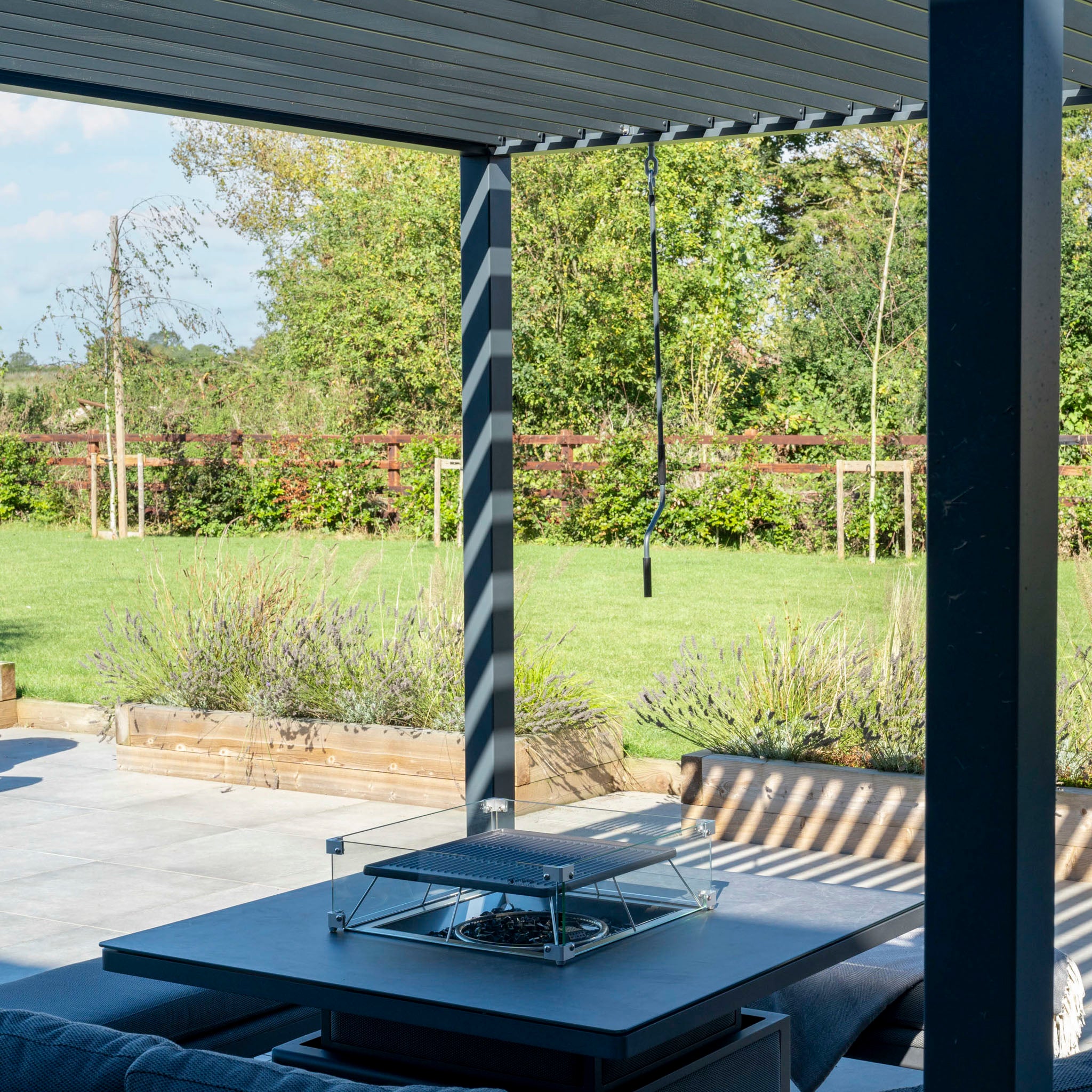 The PergoSTET Lite 3m x 3m Metal Square Pergola in Grey provides stylish shade with a waterproof roof and built-in drainage. Below, sleek furniture encircles a fire pit, all overlooking a sunlit green lawn and garden.