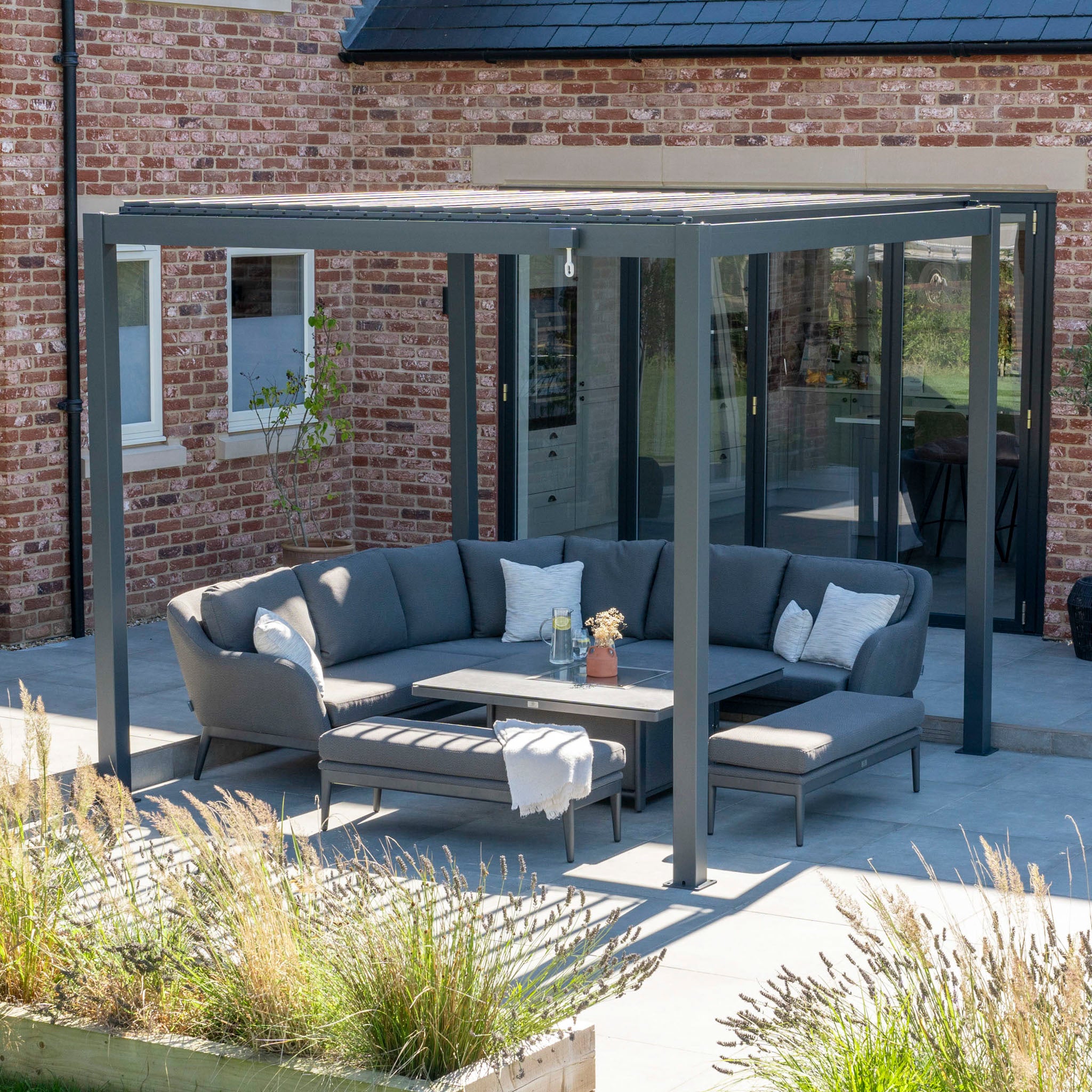 An outdoor patio features a gray sectional sofa beneath the PergoSTET Lite 3m x 3m Metal Square Pergola in Grey with a waterproof roof. A brick house provides the backdrop, while greenery adorns the foreground, crafting an inviting and stylish space.