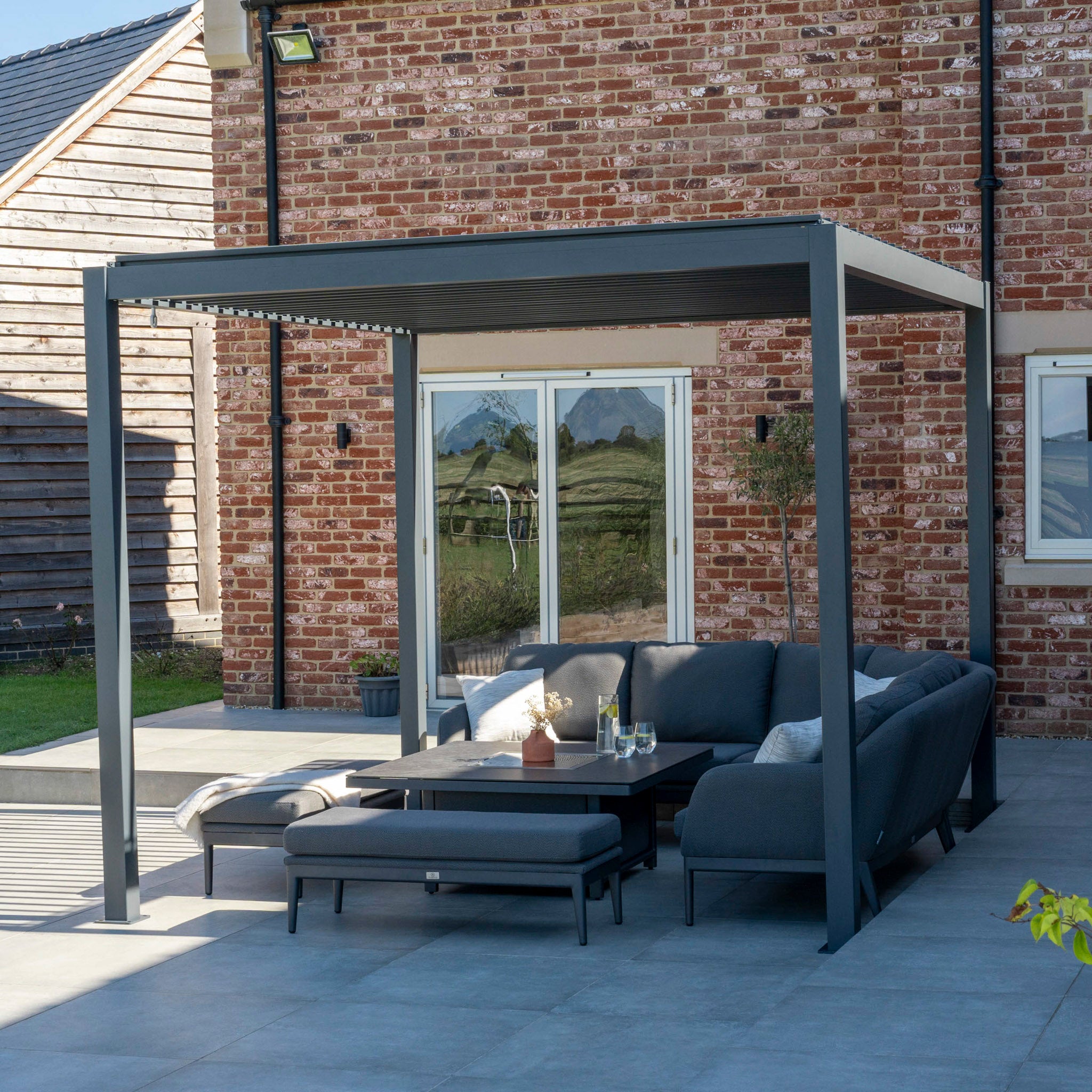 The PergoSTET Lite 3m x 3m Grey Metal Square Pergola with a waterproof roof is complemented by a gray sectional sofa and black coffee table on a tiled patio next to a brick house.