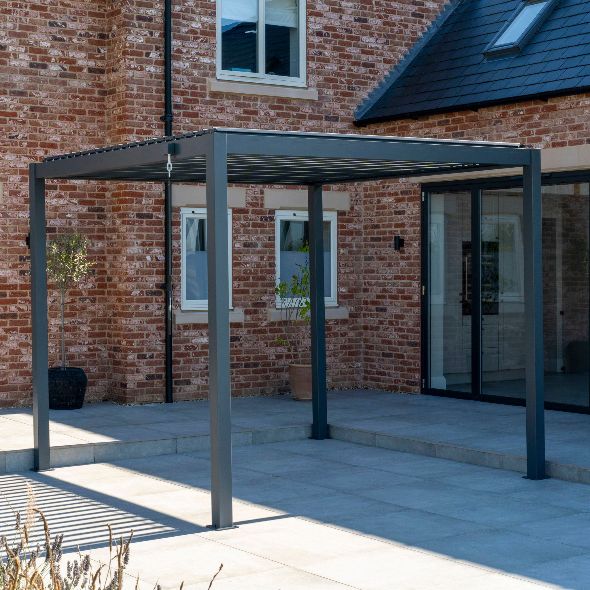The PergoSTET Lite 3m x 3m Metal Square Pergola in Grey, featuring a waterproof roof and built-in drainage system, is installed on a spacious patio outside a brick house with large windows.