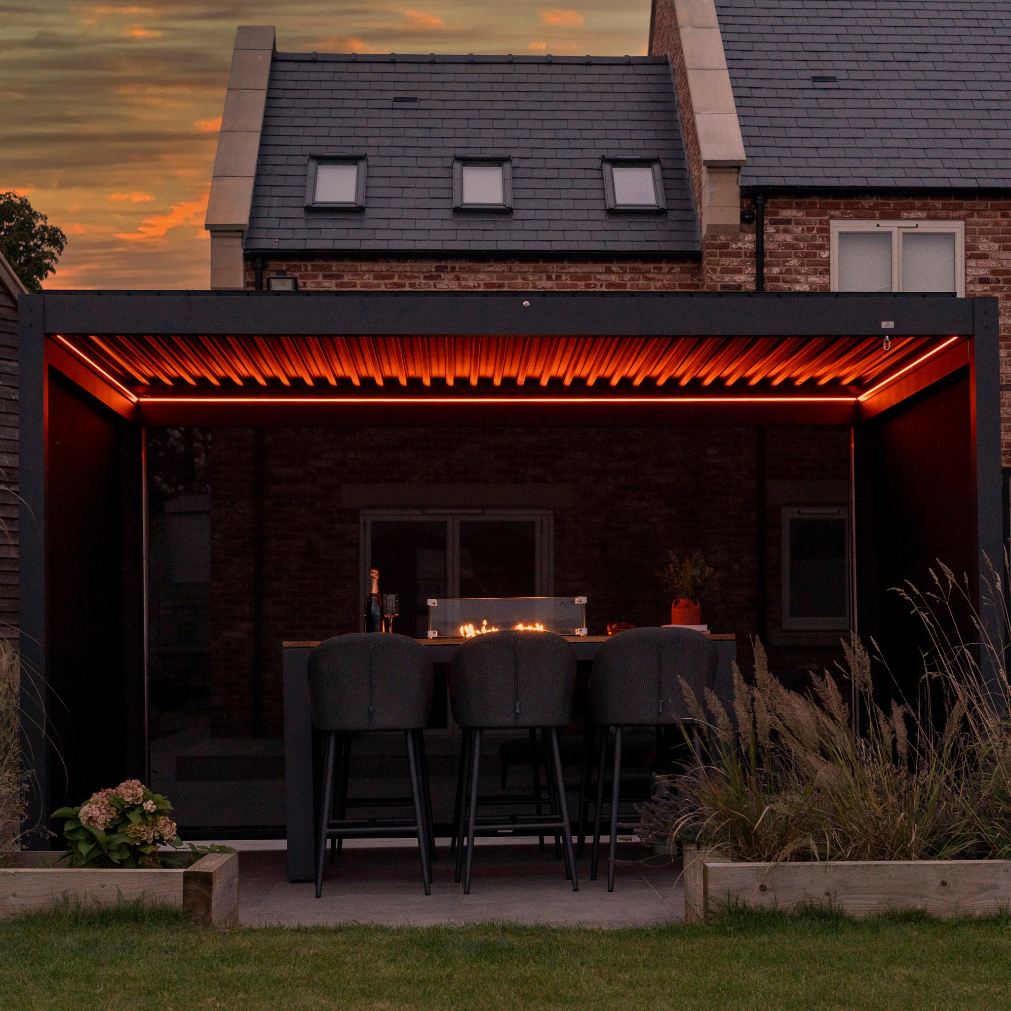 A covered outdoor seating area with black chairs and a table sits elegantly under a PergoSTET Plus 3m x 4m Rectangular Pergola with 3 Drop Sides and LED Lighting in Grey, illuminated by soft orange lights, creating a warm ambiance against the house at sunset.