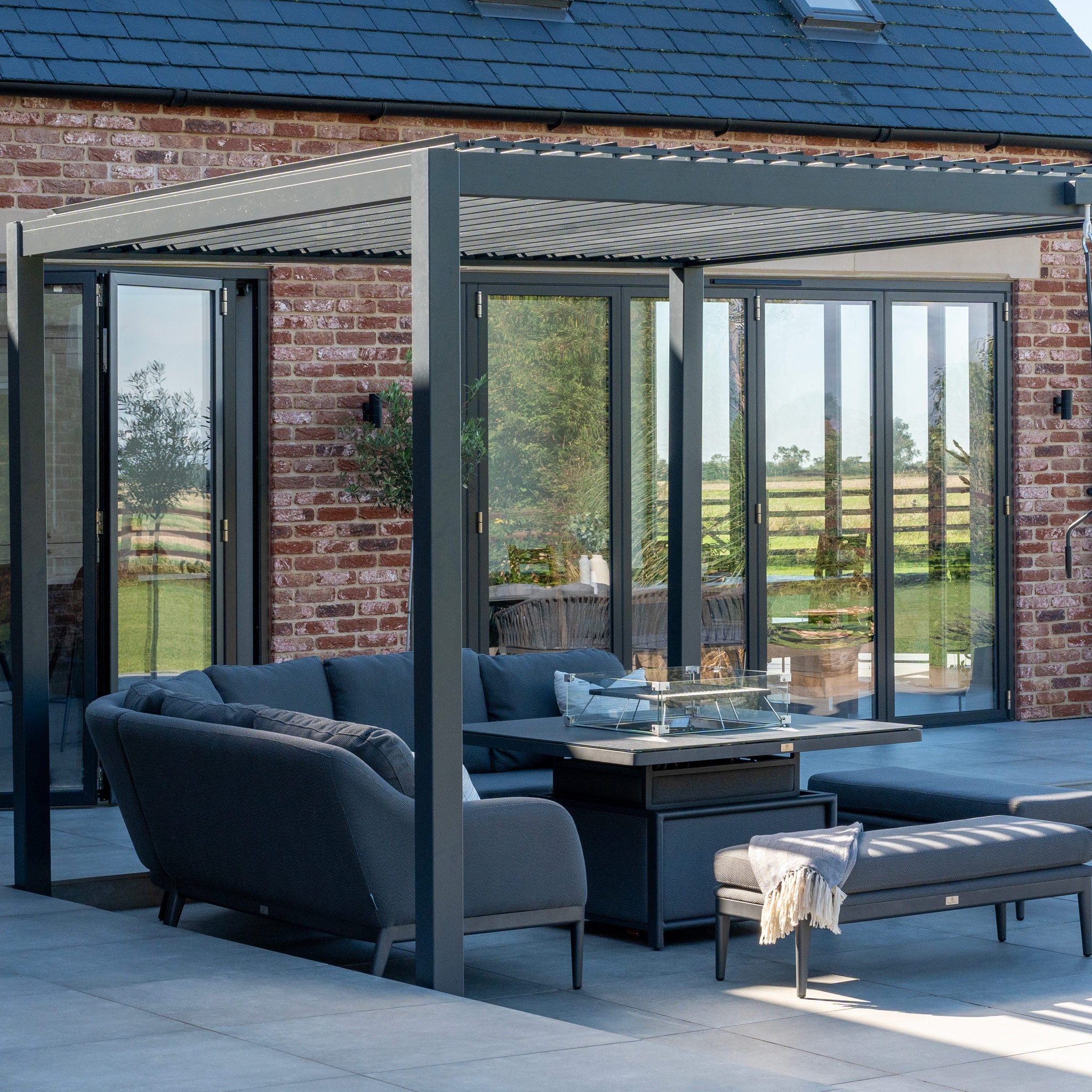 A modern patio with dark furniture lies under the PergoSTET Lite 3m x 4m metal grey pergola, featuring a built-in drainage system. Set against a brick house with large glass doors, it blends style and functionality seamlessly.