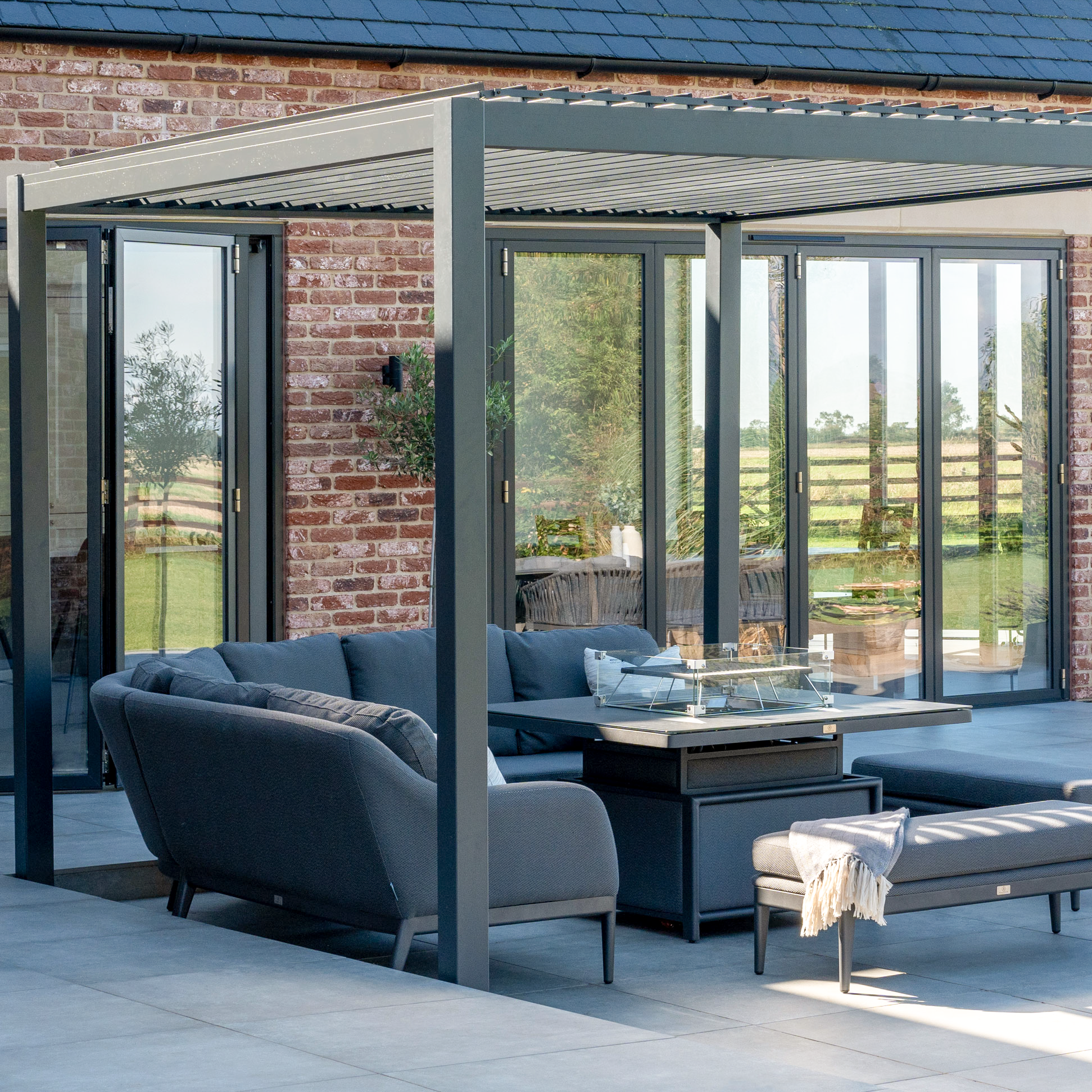 Modern outdoor patio with gray furniture, a fire pit table, and a PergoSTET Lite Neo 3m x 4m Metal Rectangular Pergola in Grey with tilting louvres beside glass doors and brick walls.