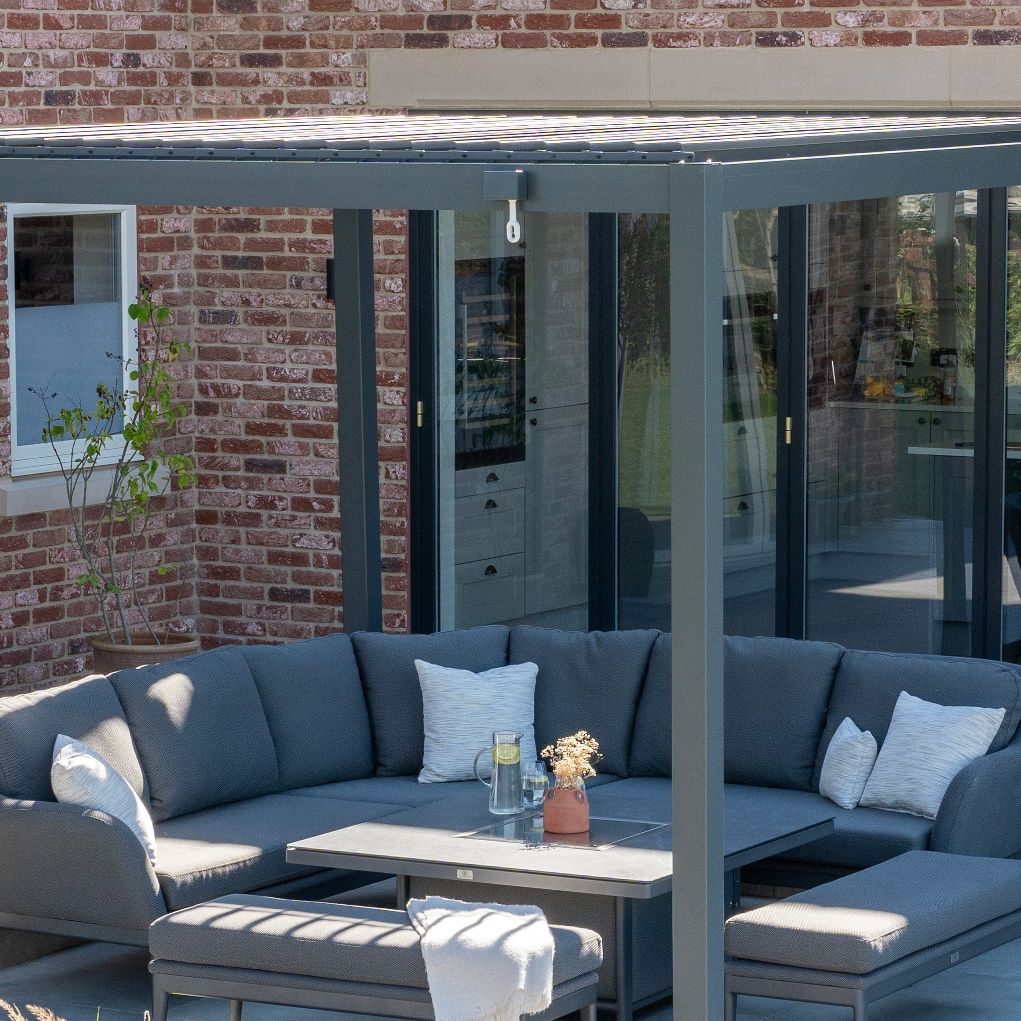 A modern patio by the brick house showcases a gray sectional sofa, pillows, and a table under a sleek PergoSTET Lite 3m x 4m Metal Rectangular Pergola in Grey.