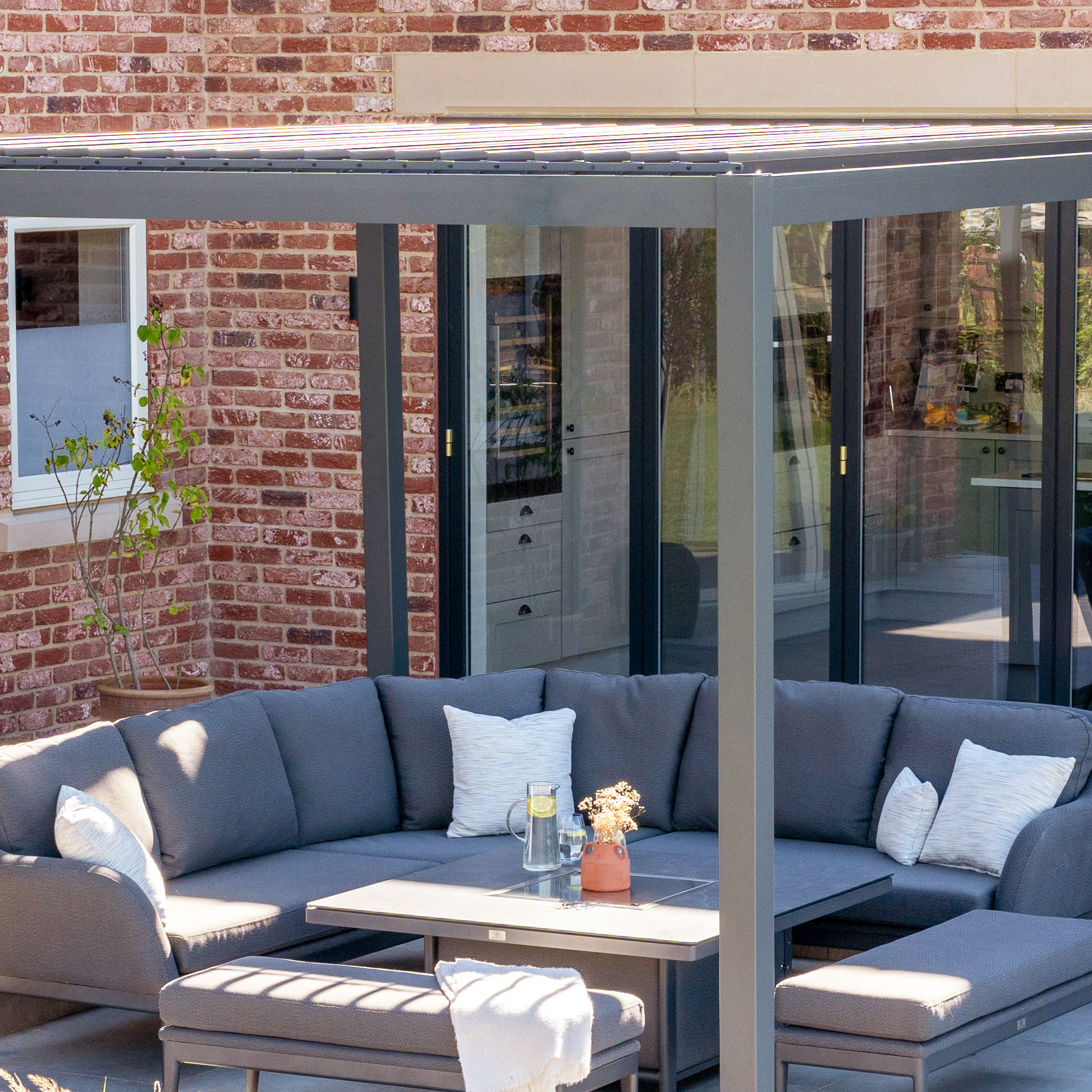 The PergoSTET Lite 2.4m x 3m Metal Rectangular Pergola in Grey stands over a gray outdoor sectional with pillows, beside a brick house and glass doors, featuring sleek tilting louvres for adjustable shade.