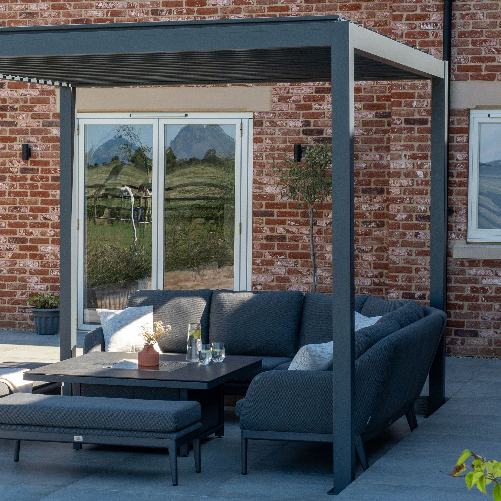 Outdoor patio featuring a dark gray L-shaped sofa, a coffee table, and a PergoSTET Lite 3m x 4m Metal Rectangular Pergola in Grey with built-in drainage in front of a brick wall and glass doors.