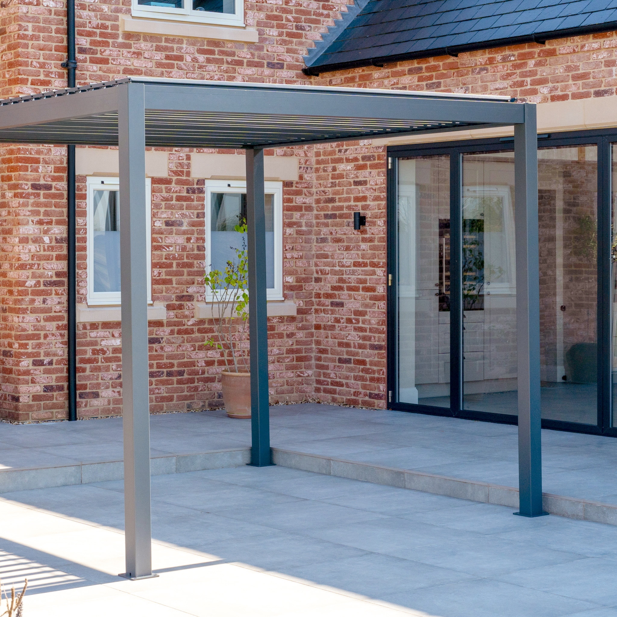 The PergoSTET Lite Neo 3m x 4m Metal Rectangular Pergola in Grey features tilting louvres and a built-in drainage system, ideal for all-weather comfort on a tiled patio beside brick homes with large glass doors and windows.