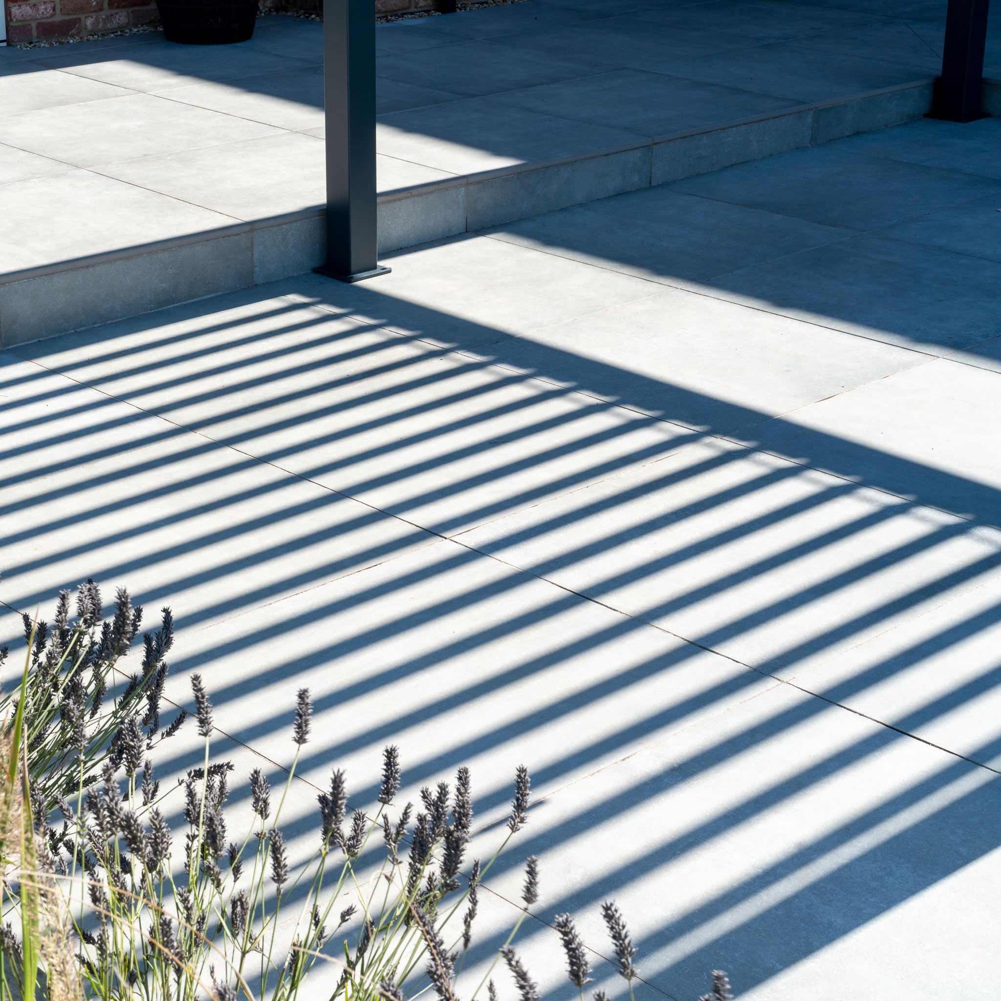 Parallel lines from a PergoSTET Lite 3m x 4m Metal Rectangular Pergola in Grey are cast on the tiled patio, accented by lavender plants in the foreground.