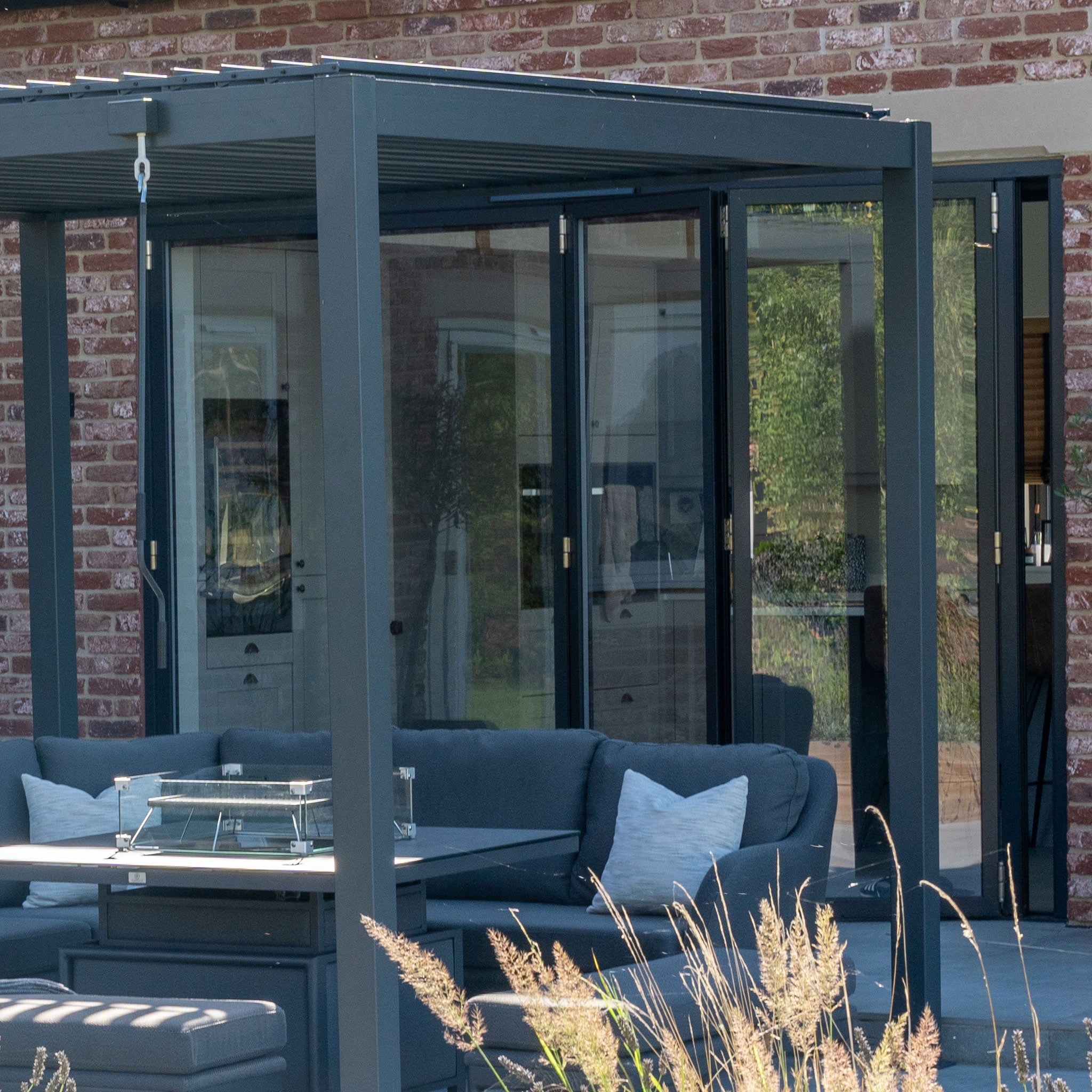 Contemporary outdoor patio with a PergoSTET Lite 3m x 4m Metal Rectangular Pergola in Grey, boasting glass panels and cushioned seating against a brick wall. The elegant design features an integrated drainage system for easy upkeep.