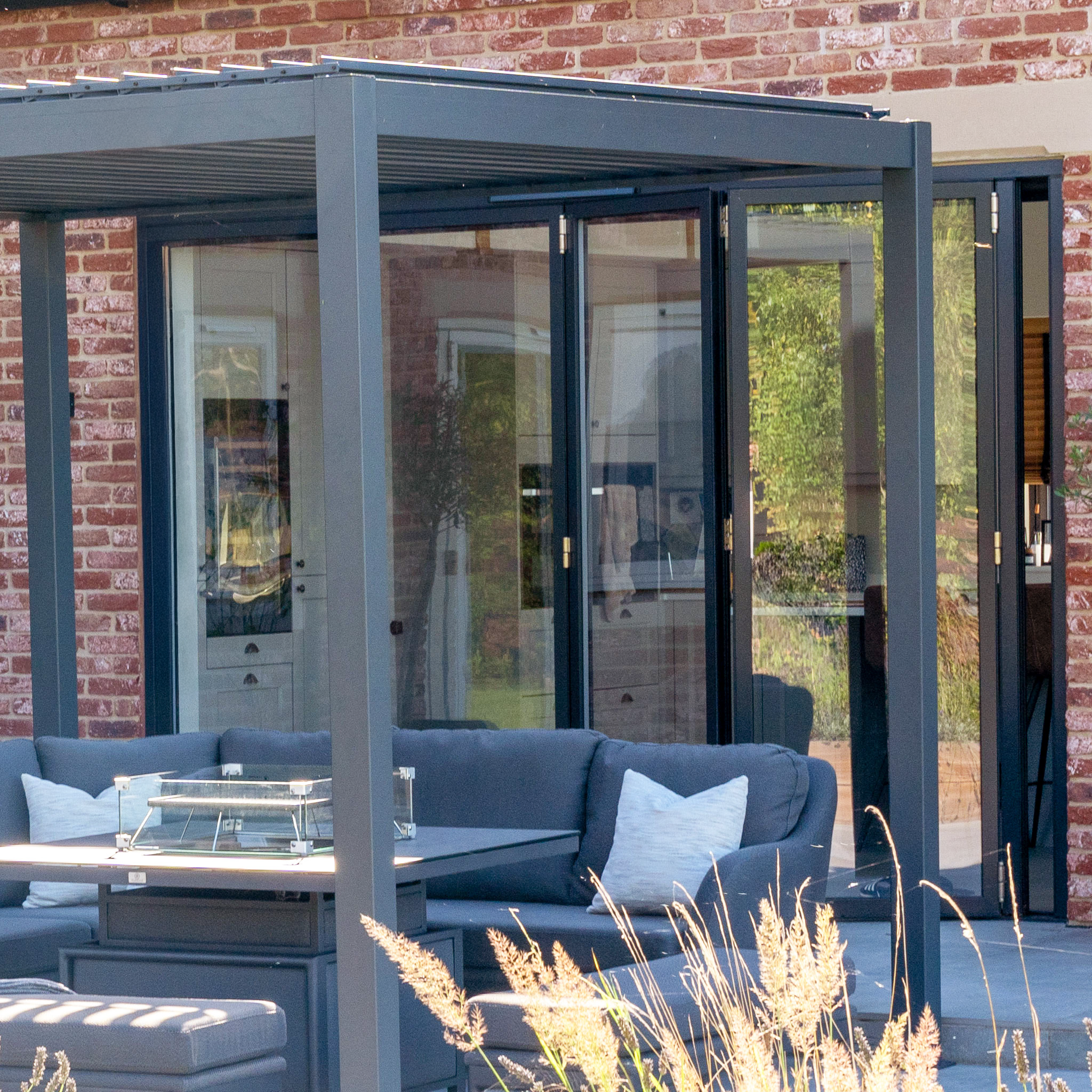Modern outdoor patio with glass walls, gray furniture, and a table beneath the PergoSTET Lite Neo 3m x 4m Metal Rectangular Pergola in Grey, featuring tilting louvres and built-in drainage, set against a brick building.