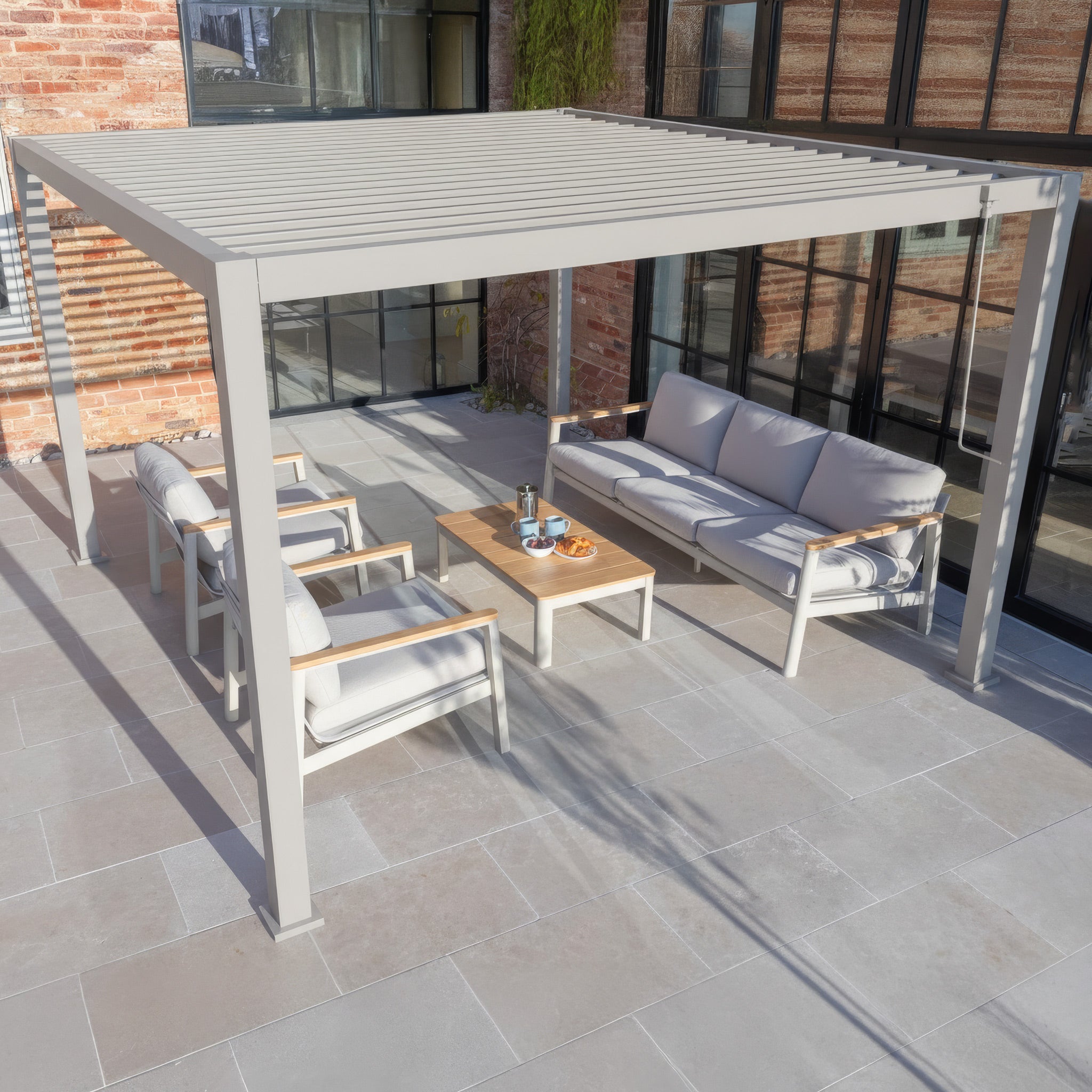 Modern outdoor patio featuring the PergoSTET Lite 3m x 3m Metal Square Pergola in Latte, cushioned seating, and a coffee table set on gray stone tiles.