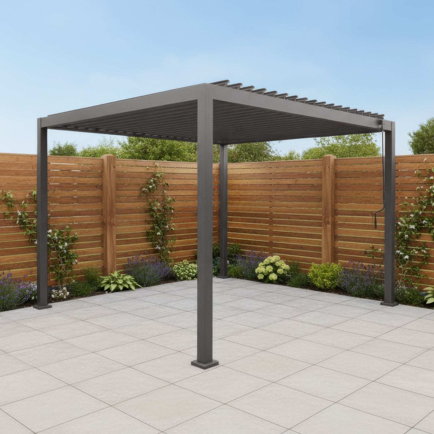 The PergoSTET Lite 3m x 3m Metal Square Pergola in Grey stands on a tiled patio, framed by a wooden fence and garden plants.