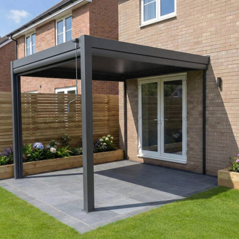 The PergoSTET Plus 3m x 3m Square Wall Mounted Pergola in grey features 3 manual side blinds, LED lighting, and a sleek built-in drainage system—perfect for patios beside sliding glass doors and wooden fencing.