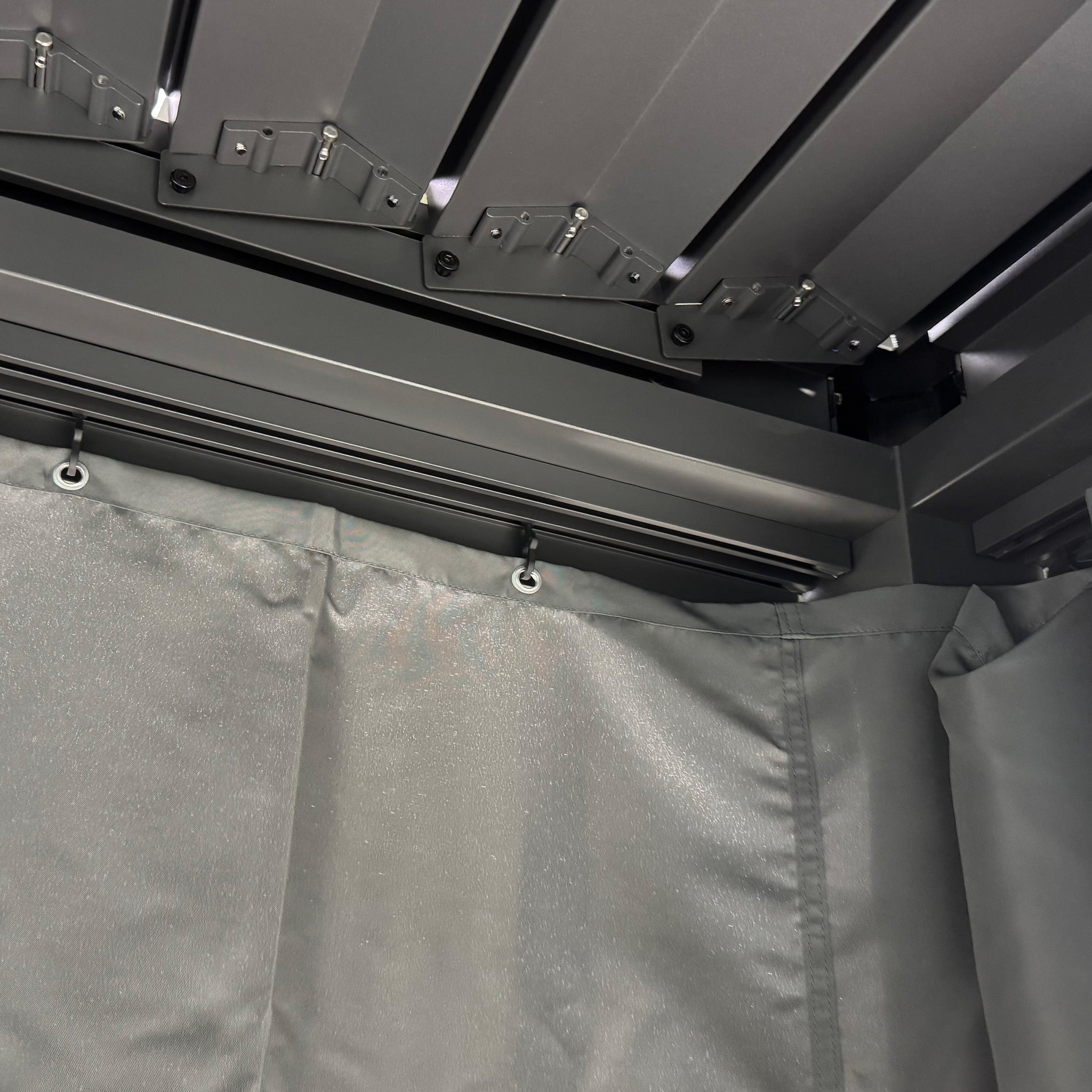 PergoSTET Side Curtains for Lite 3m x 4m Rectangular Pergola in Grey attach to a metal rail under a dark segmented ceiling, providing stylish weather protection for your pergola.
