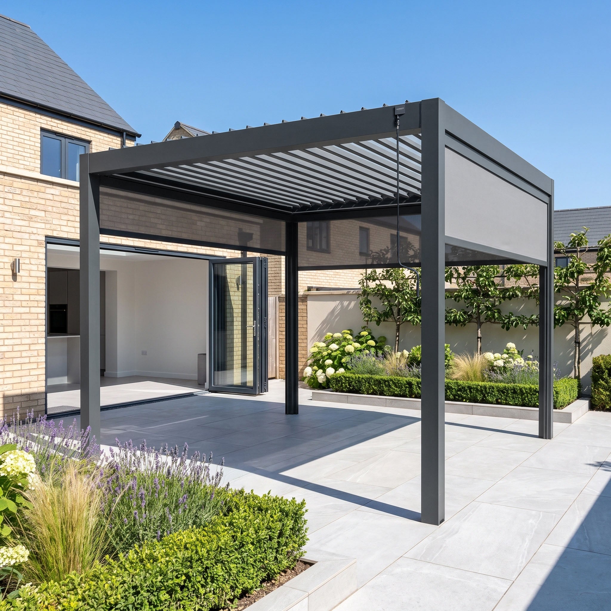 Modern patio featuring the PergoSTET Pro 3m x 3m Square Pergola in Grey, complete with three manual side blinds and integrated LED lighting. The pergola’s sleek design pairs beautifully with light tiles, lush plants, and open folding glass doors on a sunny day—delivering stylish shade for your garden.