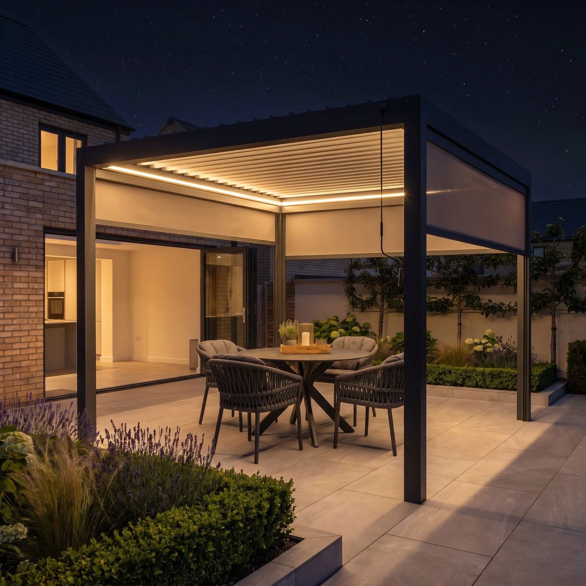 Enjoy an evening gathering in a modern outdoor patio illuminated by the PergoSTET Pro 3m x 3m Square Pergola with 3 Manual Side Blinds and LED Lighting in Grey, featuring a round table and four chairs surrounded by lush plants for elegant garden shade.